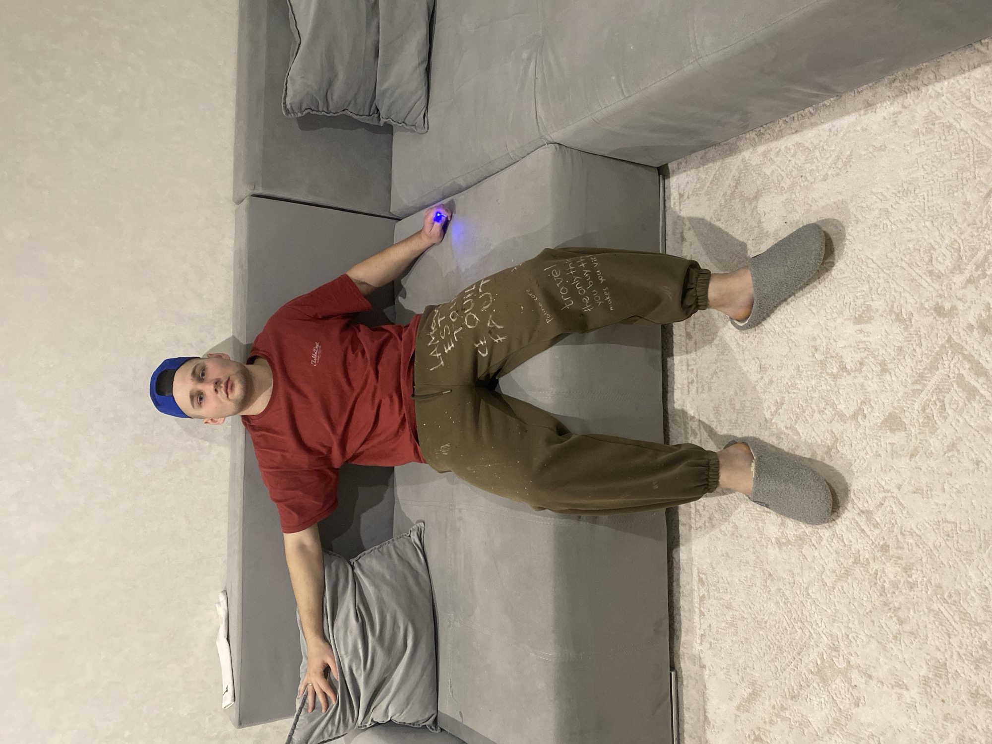 A young adult male is seated centrally on a grey fabric modular sectional sofa in an indoor environment in Chaikovsky, Russia. He wears a red t-shirt with a small dark logo on the left chest, olive green sweatpants adorned with white paint splatters and handwritten text ("LA MORT EST TOU CE QUI FAUT" on the upper left thigh, and "Polina core" above "travel the only thing you buy that makes you rich" on the lower right thigh), a blue baseball cap worn backward, and grey textured slip-on slippers with a light interior. His right arm rests on a grey throw pillow positioned on the left armrest of the sofa. His left hand, resting on the sofa cushion, holds a small, dark, cylindrical object emitting a distinct blue light. The man looks directly at the camera with a neutral expression. The background consists of a light-colored textured wall. The floor is covered by a light-colored patterned rug. A folded white item, possibly a garment or towel, is visible on the sofa backrest to the man's left.