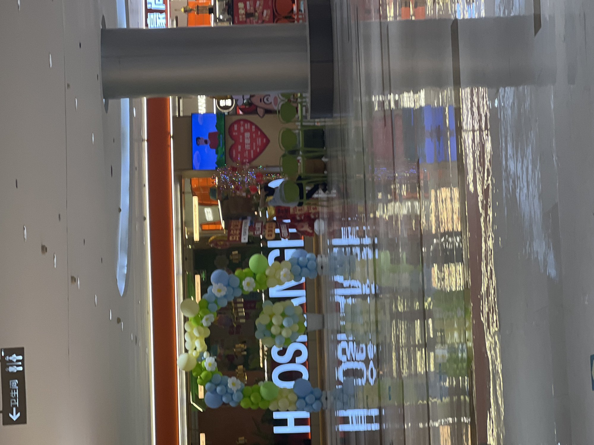 This image captures a scene inside a shopping mall in Liandu District, China. The foreground shows a wet, reflective floor with the reflections of illuminated signage and structural elements of the mall. In the middle ground, a large, illuminated sign spells out "H OSAN GH I" in white block letters, supported by a balloon arch of blue, green, and white balloons. To the right of the sign, several empty green chairs are arranged. Behind the chairs, a large red heart-shaped graphic with Chinese characters is displayed, along with a digital screen showing a cartoon character. Further in the background, a cylindrical pillar is visible, and elements of store fronts can be seen. The ceiling of the mall is visible at the top of the image, with lighting fixtures and a directional sign pointing to a restroom with male and female figures. The overall impression is of a commercial indoor space, possibly decorated for an event.