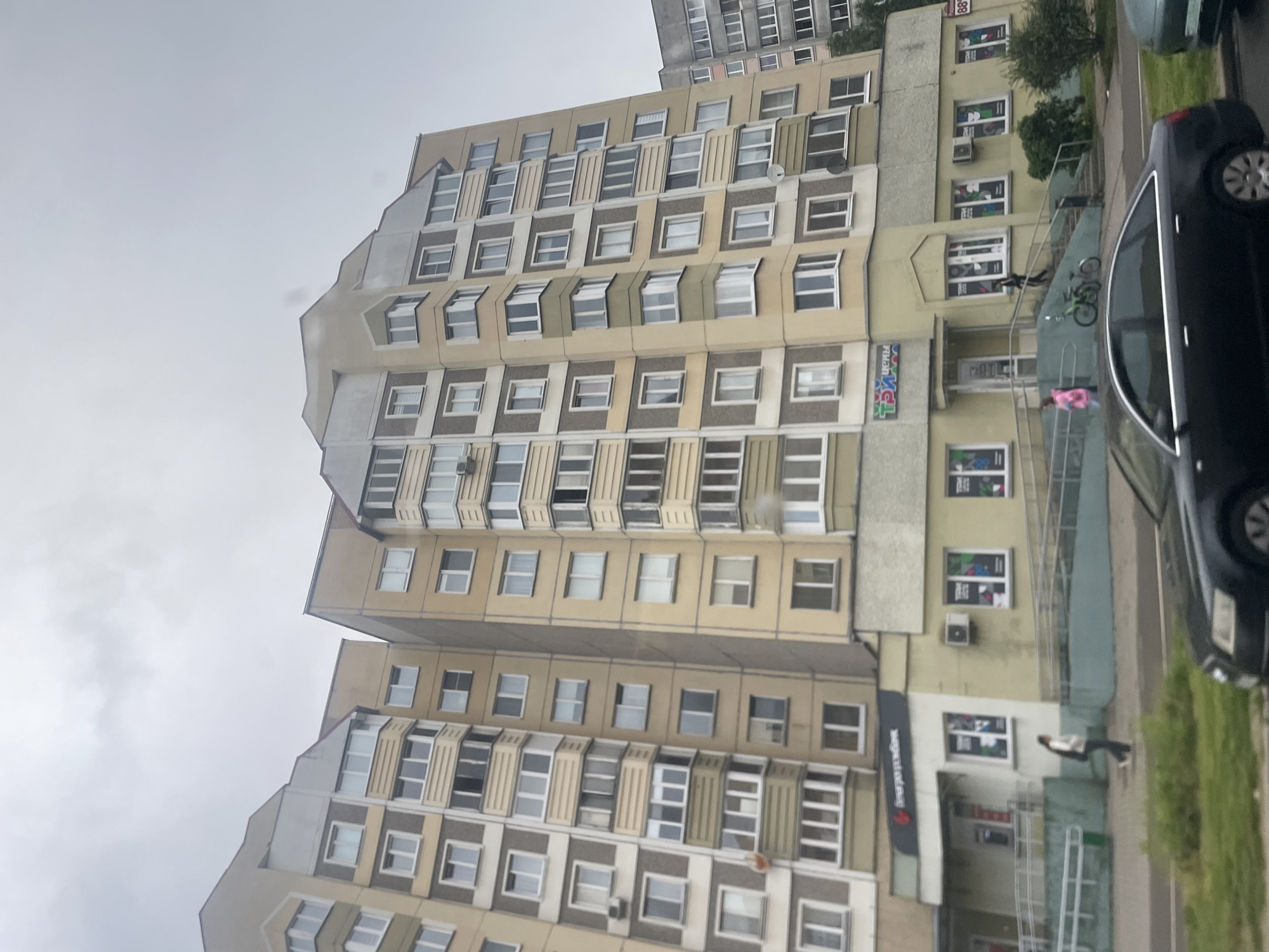 This is a medium shot, eye-level, outdoor, vertical, photographic image of a multi-story apartment building. The building is light yellow and gray, with multiple windows and balconies. In the foreground, there is a dark gray sedan and a portion of a green vehicle. A sidewalk with a ramp is in front of the building, with two people walking. One person is wearing dark pants and a white jacket, and the other is wearing a pink jacket. A green bicycle is parked on the ramp. A sign with red and white lettering is visible on the building, as well as other signs with colorful graphics. The sky is overcast and gray.