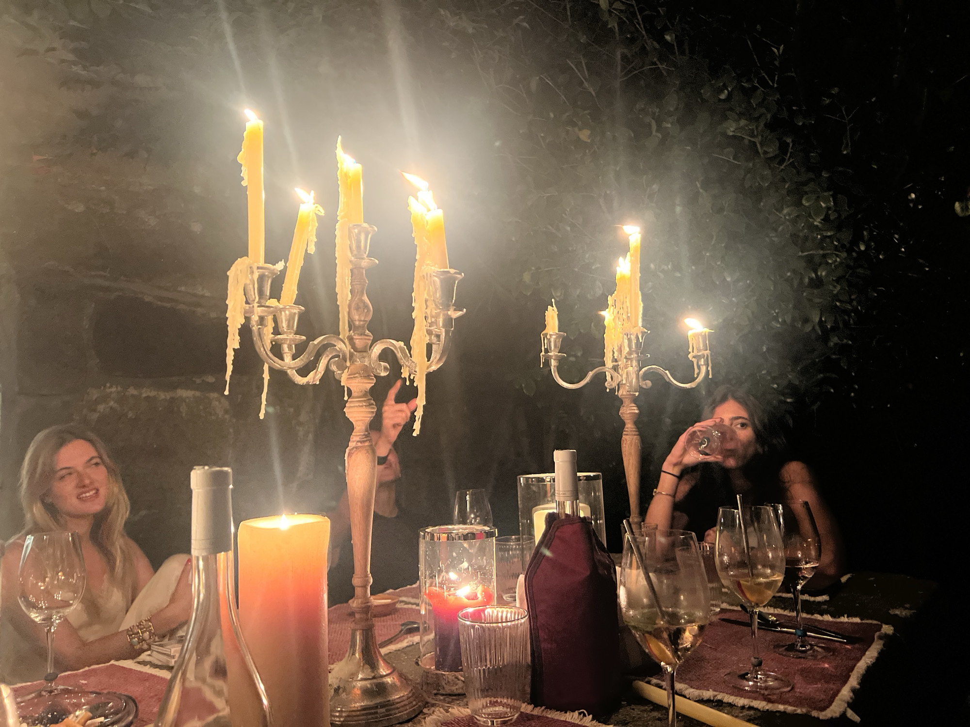 The image depicts a dimly lit outdoor dinner setting, likely in Vila Nova de Cerveira, Portugal, illuminated by several candelabras and large pillar candles. Two women are seated at a table laden with wine glasses, bottles, and a burgundy wine chiller. One woman on the left, with blonde hair, is leaning back and smiling. The other woman on the right, with dark hair, is drinking from a wine glass. In the foreground, a large orange pillar candle is lit, and partially obscured by a clear bottle with a grey cap. A silver candelabra with melting yellow candles is prominent to the left, and another smaller candelabra is visible to the right. The background consists of dark foliage and what appears to be a stone wall.