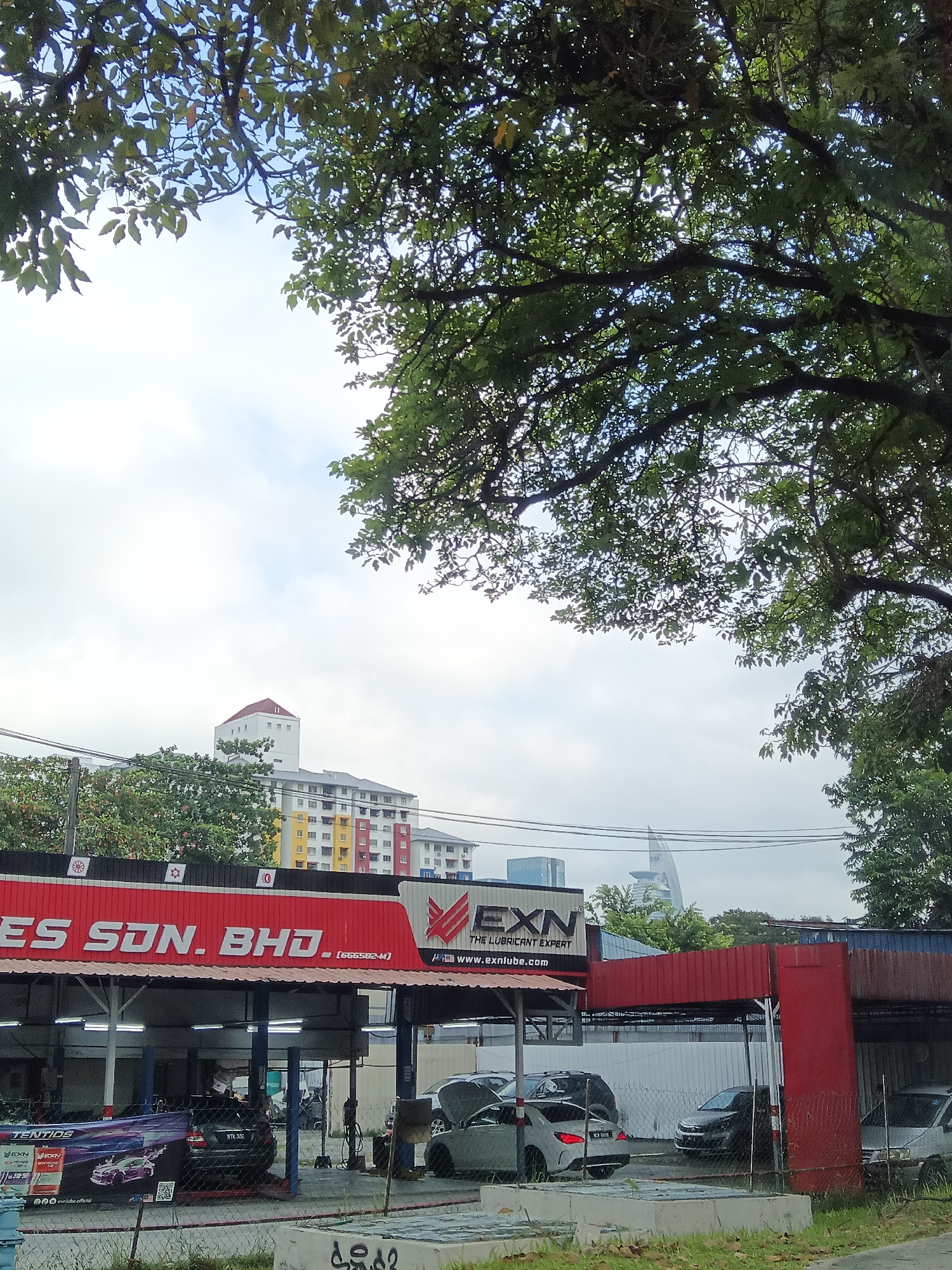 The image captures a street-level view of a car service center in Kuala Lumpur, Malaysia, under an overcast sky. Lush green tree branches frame the top and right sides of the frame. In the background, a cluster of multi-story apartment buildings with brightly colored facades (red, yellow, blue) and a tall, white building with a red-capped tower are visible. A modern, sleek skyscraper with a distinctive shape is also present in the distance.
The car service center features a prominent red awning with white lettering that reads "ES SDN. BHD." and "EXN THE LUBRICANT EXPERT." Below the awning, several cars are visible. Two cars are on lifts, suggesting maintenance is in progress. One dark-colored sedan is parked on the ground to the left, and a white sedan is parked in the middle of the lot. Another car is partially obscured on the right. A large banner on the left advertises "TENTIOS" and "EXN" products with images of a purple car. Electrical wires are strung across the scene, and a chain-link fence runs along the front of the service area. In the foreground, there are concrete slabs and grassy areas.