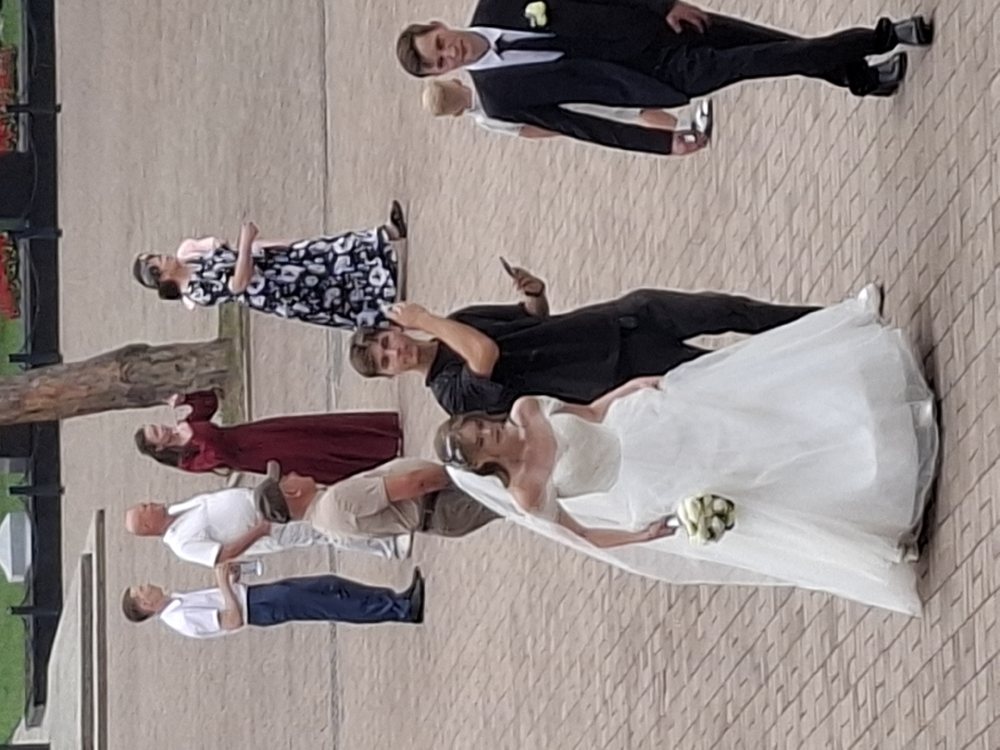 The image depicts an outdoor scene, likely a public square or plaza, with cobblestone paving. Several individuals are visible, suggesting a gathering or event.
In the foreground, a woman in a white wedding dress with off-the-shoulder sleeves and a veil is walking from left to right. She is holding a bouquet of white flowers. To her right and slightly behind her, a young man in a black t-shirt is looking towards the camera, holding a mobile phone in his right hand and what appears to be another device in his left. Further to the right and behind him, a man in a dark suit with a white shirt, black tie, and a boutonnière is walking towards the camera. A young boy in a light-colored shirt is visible to the left of the suited man.
In the background, on the left side of the image, a man in a white shirt and dark trousers stands next to another man in a light-colored polo shirt and khaki trousers. A woman in a dark red, floor-length dress is standing behind them, gesturing with her right hand. Further to the right and behind this group, a woman in a black and white patterned dress is visible.
The environment includes a paved area, hints of greenery and structures in the upper background, and a tree trunk on the right. No specific landmarks indicating Almaty, Kazakhstan are visible in the image.