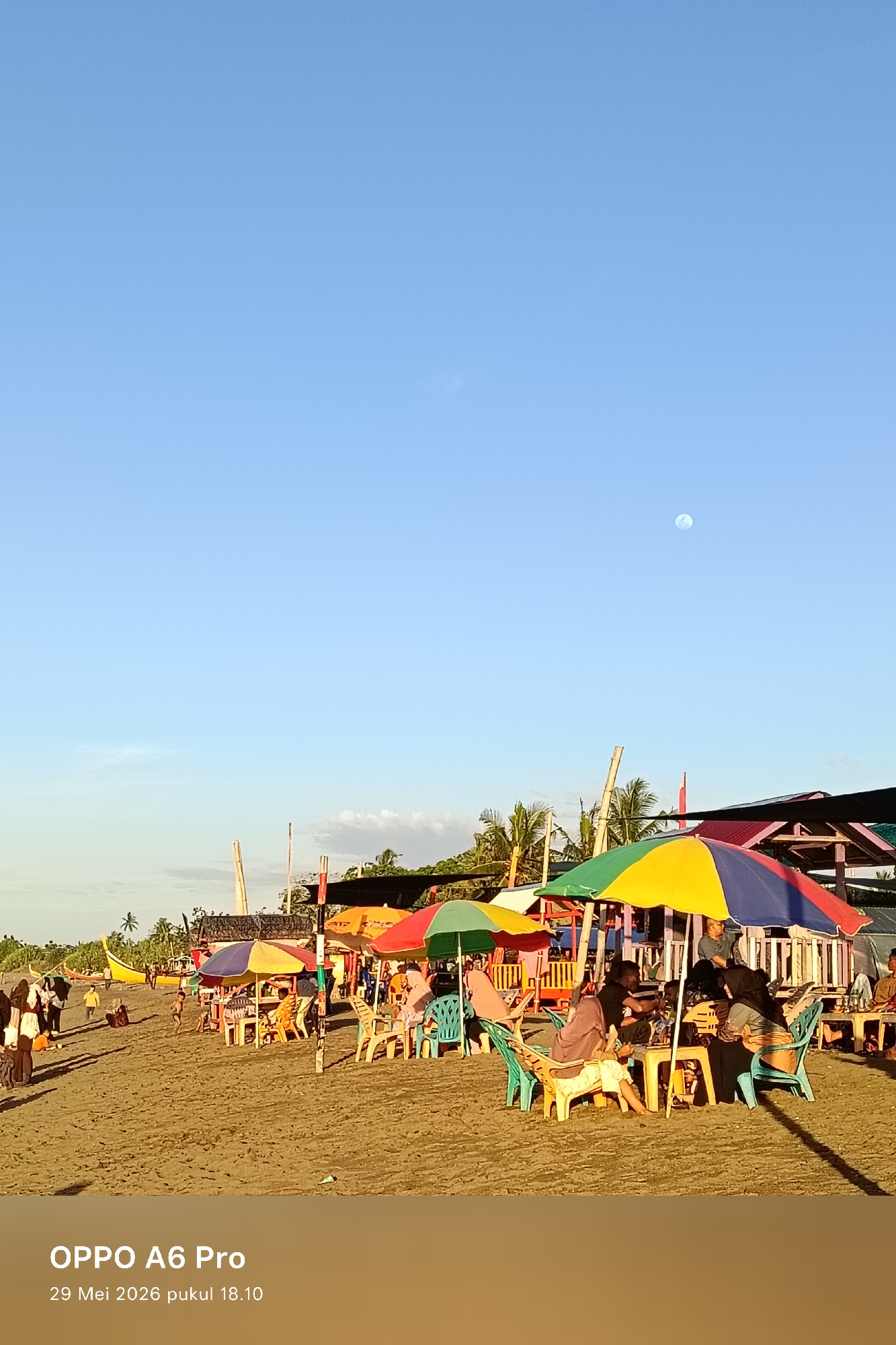 The image depicts a beach scene in Rheumbaroh, Indonesia. Several colorful umbrellas provide shade for seated individuals at tables. People are walking along the beach in the background, and a boat is visible. The sky is clear and blue with a small, pale moon visible high above. The timestamp indicates the photo was taken on May 29, 2026, at 18:10.