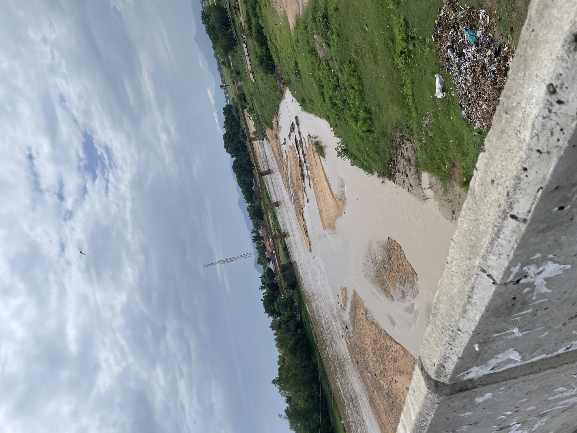 The image depicts a wide, muddy river flowing through a landscape of green vegetation and scattered buildings under a cloudy sky. In the foreground, a concrete barrier, possibly part of a bridge or embankment, is visible, with some peeling white paper adhered to its surface. The river itself has exposed sandy banks and areas of shallow water. Across the river, a line of trees forms a natural border, behind which are some houses with red roofs and a tall communication tower. A bridge, constructed of concrete pillars and a flat deck, spans the river in the middle ground. Beyond the trees and buildings, a range of low mountains can be seen in the distance. The right side of the image shows a grassy embankment with a pile of garbage, including plastic bags, bottles, and debris. A single, distant kite shaped like a dragonfly is visible in the sky. The overall impression is one of a rural or semi-urban setting, possibly Jalingo, Nigeria, with evidence of both natural elements and human activity.
