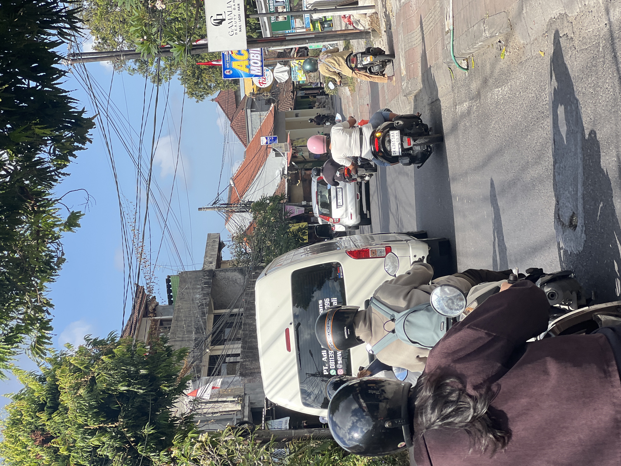 The image depicts a street scene in Denpasar, Indonesia, characterized by a moderate amount of traffic, primarily motorcycles. The time of day appears to be daytime, with bright sunlight casting shadows. In the foreground, two individuals are riding a motorcycle, both wearing helmets. The rider in the back is wearing a maroon jacket and has long dark hair, while the rider in front is wearing a light-colored hoodie and a light blue backpack.
To their right, another motorcycle is visible with a rider wearing a light pink helmet and a white shirt. Further down the street, more motorcycles and a white van are present. The van has text on its rear window, including what appears to be a company name and contact information.
The environment features buildings lining the street, with traditional Indonesian architecture including red-tiled roofs. Numerous electrical wires are strung across the sky, a common sight in urban Indonesian settings. Signs are visible on the buildings, some advertising businesses. The road surface is asphalt, with a paved sidewalk on the right side of the street. Vegetation, including trees with green foliage, can be seen on both sides of the road and on top of some of the buildings. The overall impression is of a busy, everyday street in an Indonesian city.