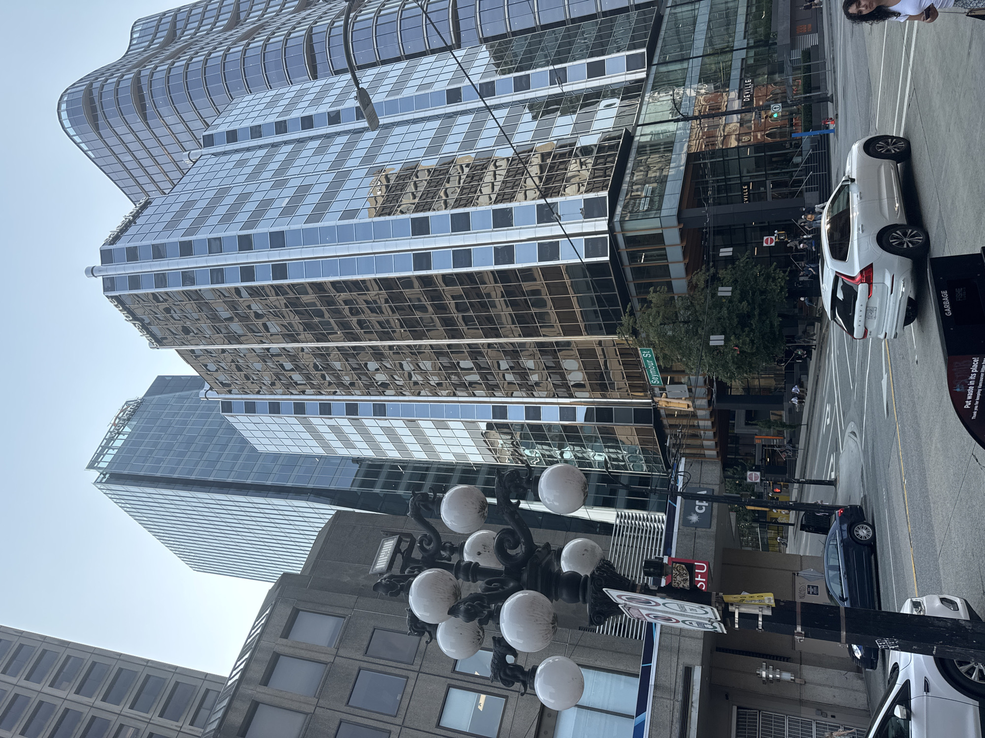 This image depicts a street scene in downtown Vancouver, Canada, likely near the intersection of Seymour Street and West Georgia Street. Multiple modern glass-and-steel office buildings dominate the skyline, their reflective facades mirroring the sky and surrounding architecture. One prominent building displays the "Salesforce" logo. In the foreground, a vintage-style street lamp with multiple globes stands beside a traffic light and various street signs. A white Volvo SUV is driving on the street, and other vehicles are parked or moving in the background. Pedestrians are visible on the sidewalks and crossing the street. The environment appears to be an urban commercial district with clear skies.