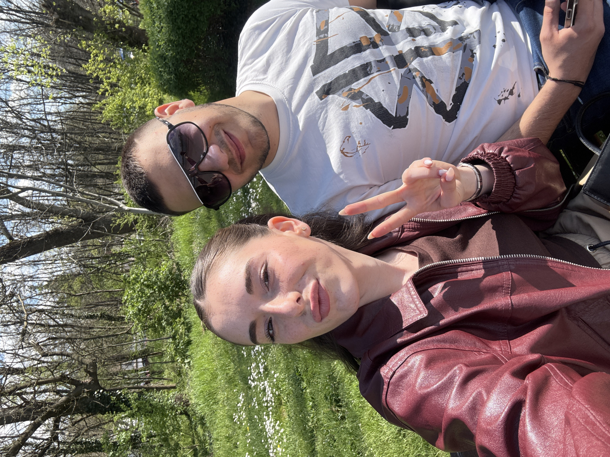 Two individuals, a male and a female, are posing for a photograph outdoors. The male is wearing sunglasses and a white t-shirt with a graphic design. The female is wearing a maroon leather jacket and making a peace sign with her fingers. The setting appears to be a grassy area with trees in the background. The location is Plovdiv, Bulgaria.