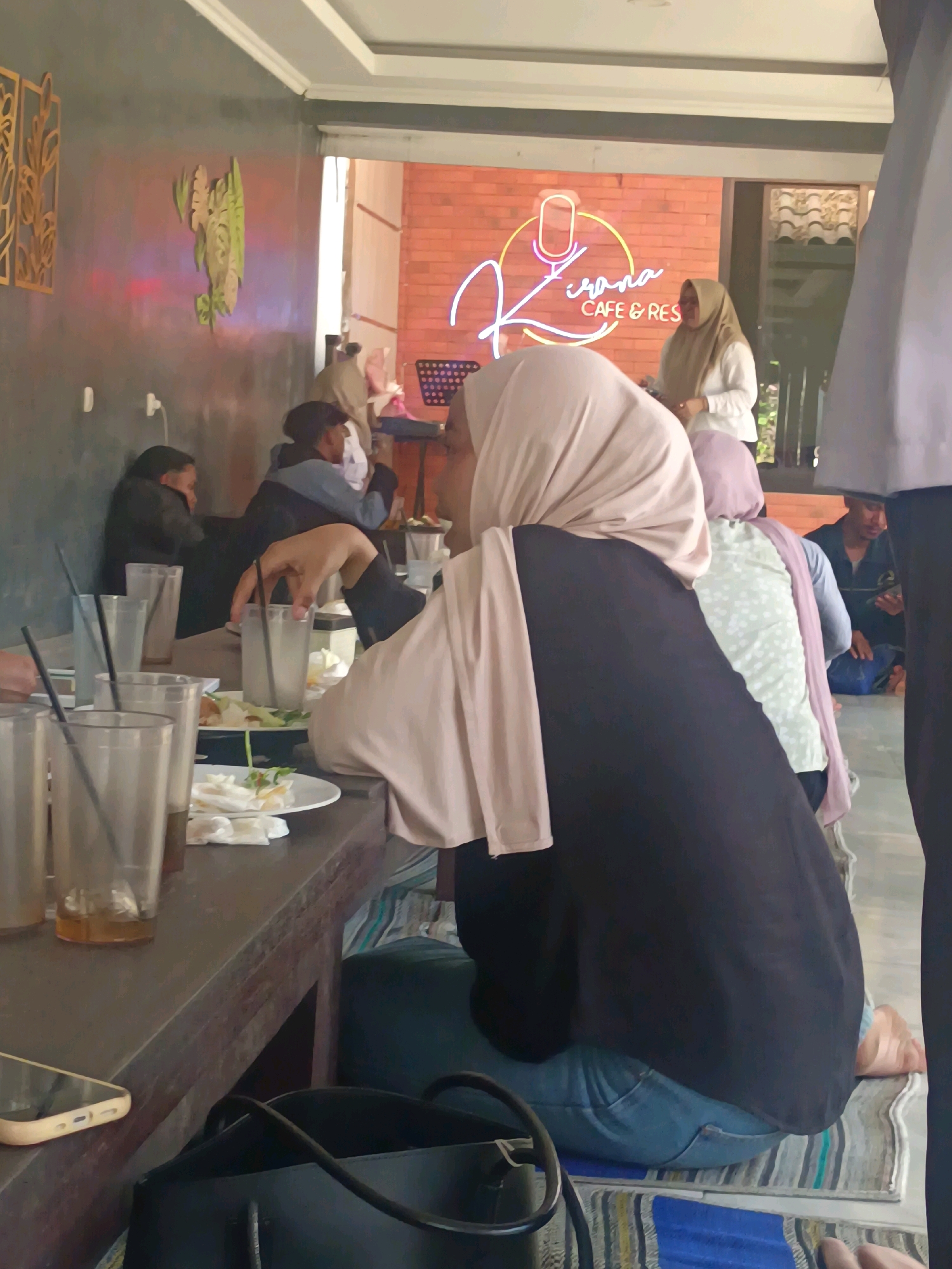 This image depicts the interior of "Kirana Cafe & Rest" in Kerangkulon, Indonesia. The scene is set at a low wooden table where several individuals are seated, engaged in dining. In the foreground, a woman wearing a light brown hijab and a dark long-sleeved top is seated, with her back partially to the camera, facing towards the table. Her left hand is visible, holding a glass of what appears to be iced tea with a straw. Multiple glasses, some with straws, and plates with food and napkins are on the table.
To the left of the foreground woman, another person in a dark jacket is partially visible. Behind them, on the wall, a stylized painting of green foliage with subtle red and yellow lighting effects is displayed. Further back, two more individuals are seated at the table, one wearing a light colored hijab and the other a blue jacket and dark head covering.
The background reveals the cafe's branding with a neon sign that reads "Kirana Cafe & Rest" in cursive script, set against a brick wall. A person in a light colored hijab and white top is standing near the sign, possibly an employee. The overall environment appears casual and serves as a communal dining space.