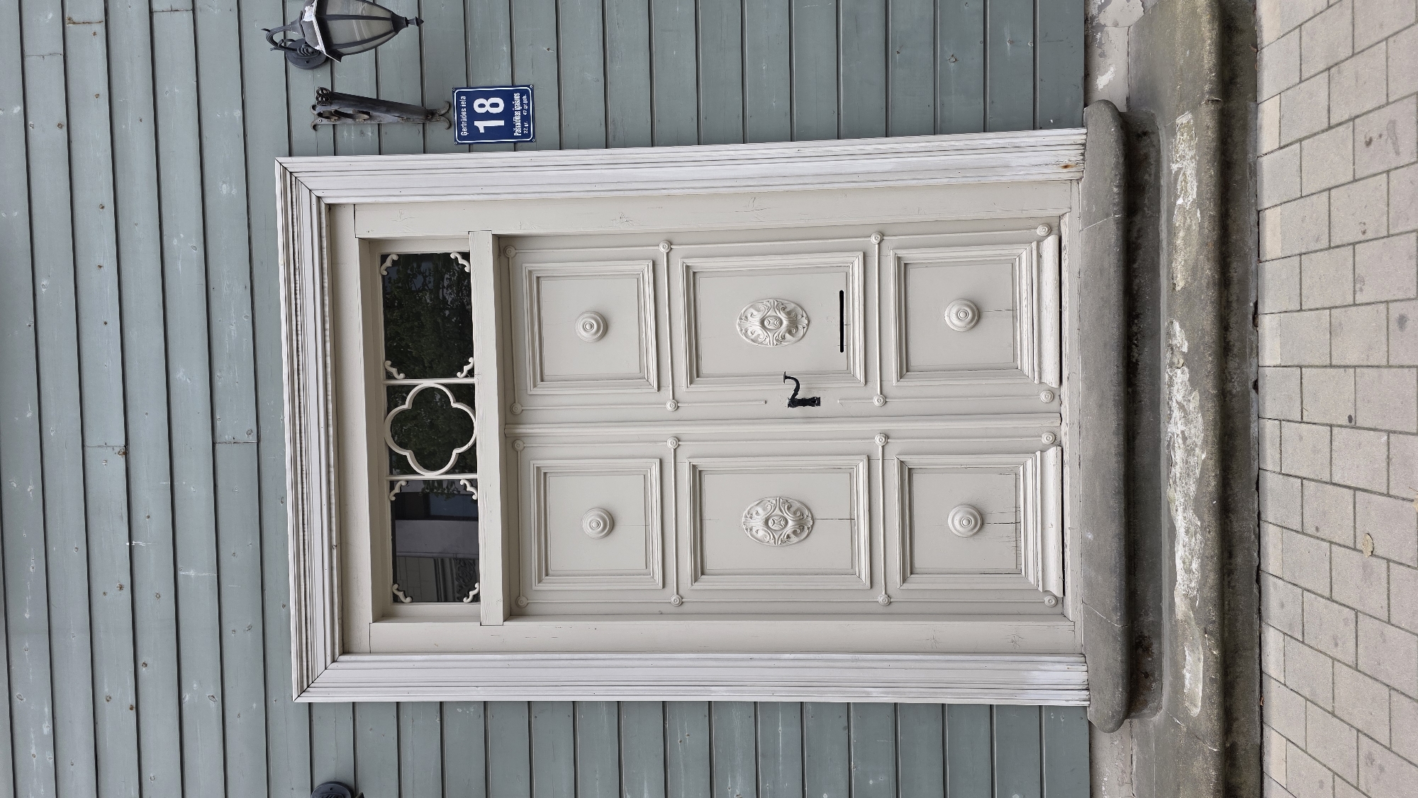 The image shows a light-colored double door with ornate panels and a small window with a decorative element above it. The door is set within a white frame and is accessed by two stone steps. The building exterior is clad in horizontally laid, pale green wooden planks. To the right of the door, mounted on the wall, is a dark-colored lantern and a blue street sign displaying the number "18" and text in Latvian. The foreground is paved with light-colored rectangular bricks. The scene appears to be a building entrance in a city setting, likely Riga, Latvia, given the Latvian text on the sign.