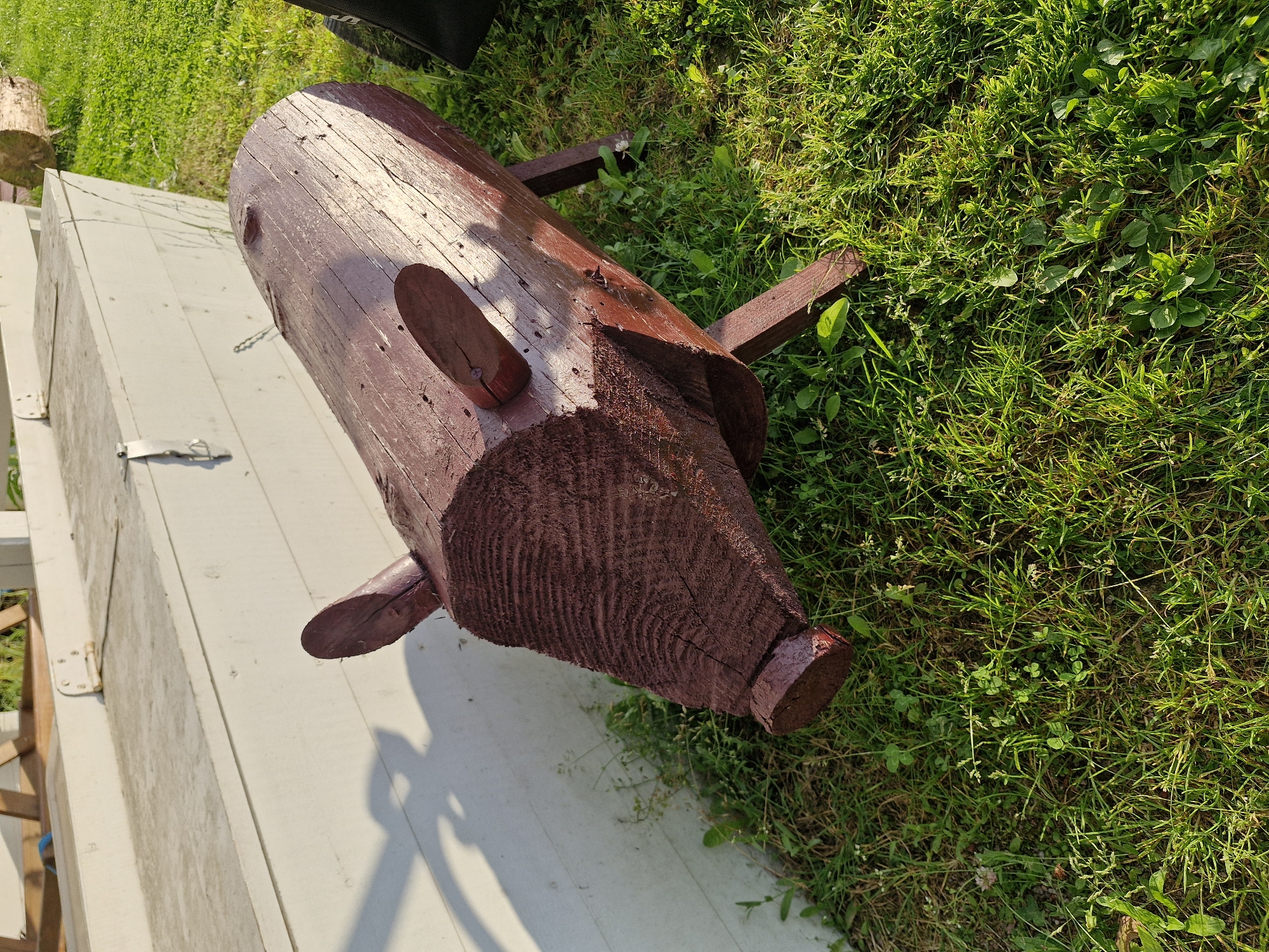 A wooden log carved to resemble a pig, painted dark red, rests on a grassy area. The pig has two small, flat ears and a snout. Its body is cylindrical with visible wood grain. Four wooden legs support the body. Behind the pig is a white, flat-topped structure, possibly a bench or a boat, with a shadow cast on it from an unseen object. The area is illuminated by sunlight, creating distinct shadows. The ground is covered in green grass and small plants. The location is the city of Sorochkino, Russia. There are no people or animals visible in the image.