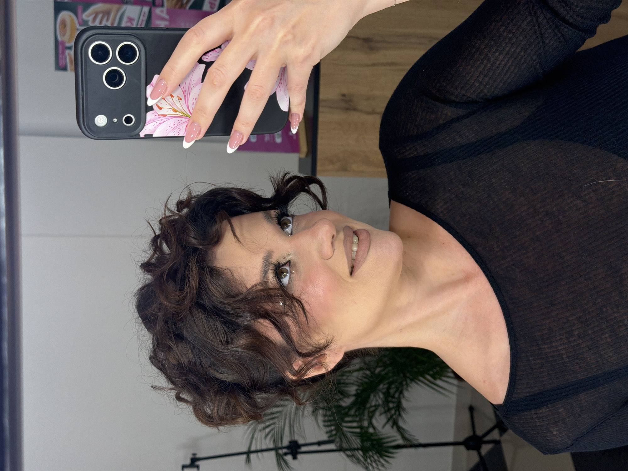 A woman with curly dark brown hair and fair skin is holding a black smartphone with a pink floral design on the back. She is wearing a black, slightly see-through, long-sleeved shirt. Her nails are manicured with a French tip design. The background is blurred, but there's evidence of a light-colored wall, what appears to be a mirror, and possibly some decorations. The image appears to be taken indoors. There is no information to suggest this image was taken in Belgrade, Serbia.