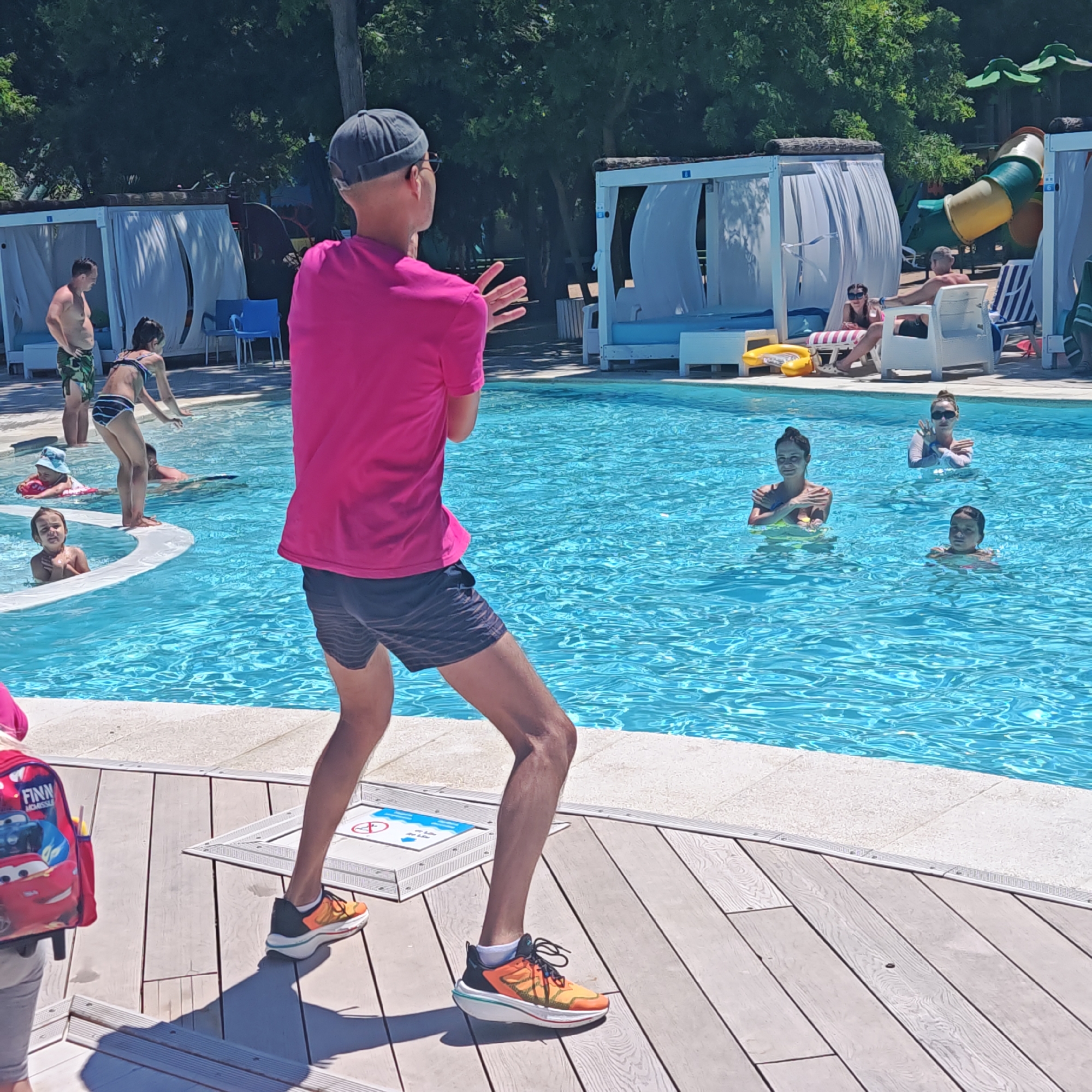 An overhead shot captures a group of people in and around a swimming pool on a sunny day. In the foreground, a person wearing a pink t-shirt, dark shorts, and bright orange and yellow sneakers stands on a wooden deck, facing away from the camera and towards the pool. Their arms are raised, hands outstretched, suggesting they are leading an activity. To the left, a child's backpack with a "Finn" label and a Cars movie design is visible on the deck.
In the pool, several individuals are engaged in various activities. Near the edge, a child in a blue hat floats on a pink ring, while another child, partially submerged, looks towards the viewer. Further into the pool, a person in a black and white striped bikini stands with their hands reaching down. To the right, three women are in the water, with two of them in the middle of the frame striking poses with their arms crossed over their chests. One woman in the background, wearing sunglasses, also has her arms crossed.
The pool is surrounded by a light-colored deck and a stone border. In the background, white cabanas with curtains line the pool area, and lush green trees form a dense canopy. A yellow inflatable ring is visible near one of the cabanas, and a colorful slide can be seen in the upper right corner. Sunlight casts shadows on the wooden deck.