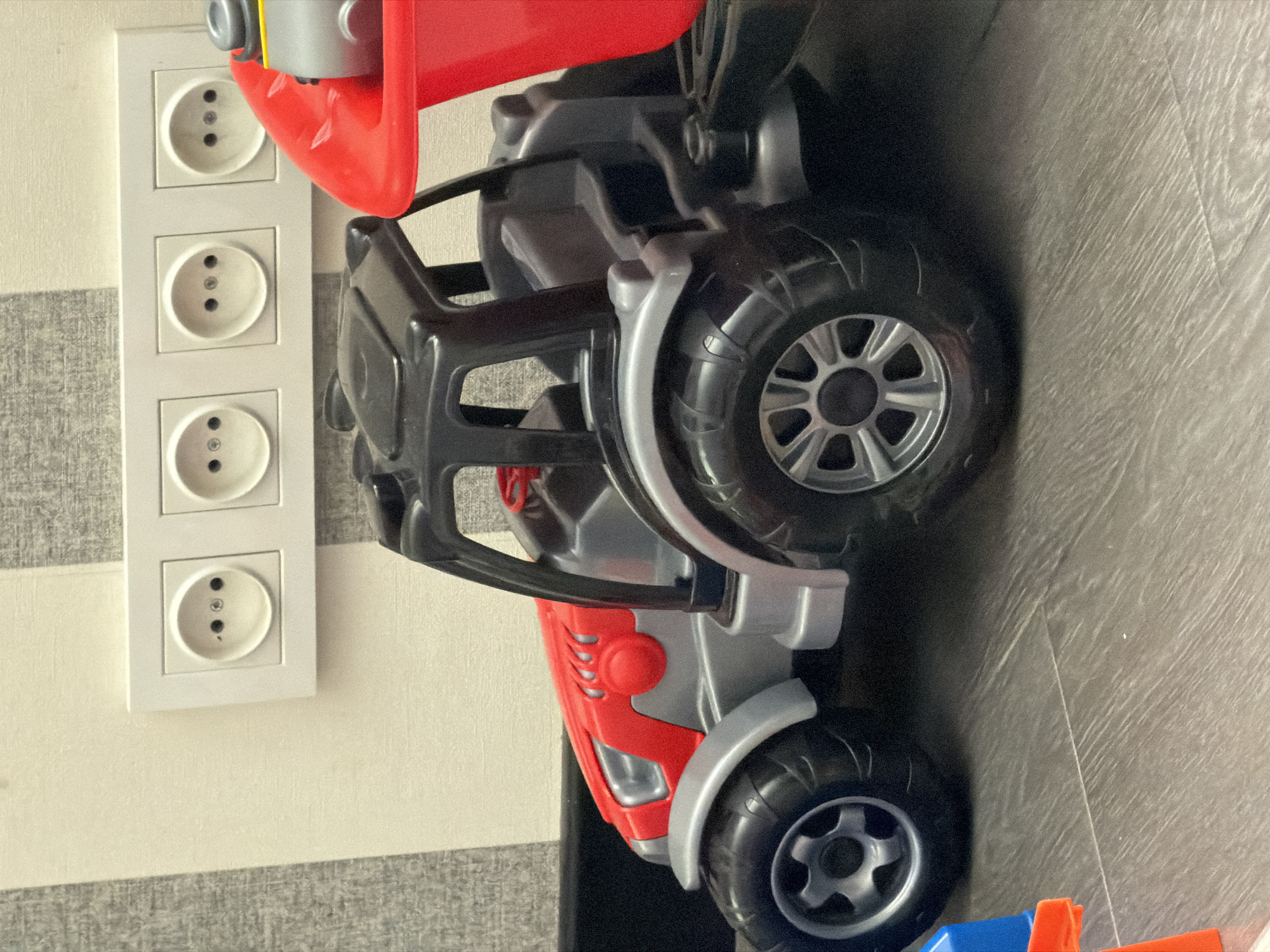 The image shows a close-up, side-view of a red and grey toy truck positioned on a floor with a striped wallpapered wall behind it. The truck has large, black, ridged tires with silver rims. The body of the truck is primarily red and silver, with black plastic detailing.
On the wall, there are three white electrical outlets stacked vertically. The outlets appear to be of a European type, with two round openings and a central grounding pin. The wall has alternating horizontal stripes of a light beige color and a textured grey pattern.
The lighting suggests an indoor setting, likely during the daytime due to the brightness. The focus is on the toy and the electrical outlets, with no people visible. The overall scene is that of a child's toy placed near a wall with electrical fixtures. There is no visible text in the image. The location context provided is Bila Tserkva, Ukraine.
