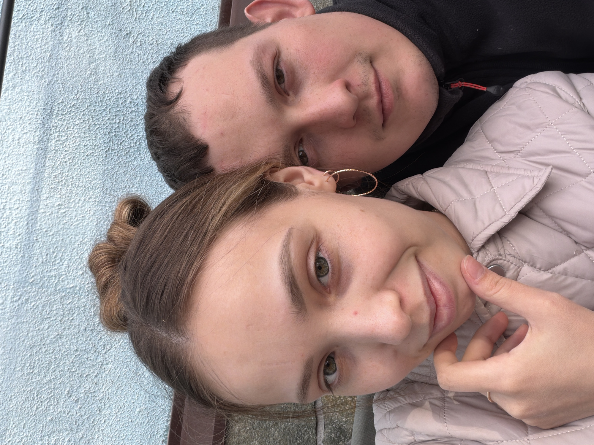 A selfie shows a man and a woman, presumed to be a couple. The man is positioned above the woman, with their heads close together. He has short, dark hair and fair skin, and is wearing a black hooded sweatshirt. The woman has her hair pulled up in a bun and is wearing a light-colored quilted jacket. Her hand is near her face. A gold hoop earring is visible on her left ear. They are outside, with a light blue textured wall in the background. The location is potentially in Прикамский, Russia.