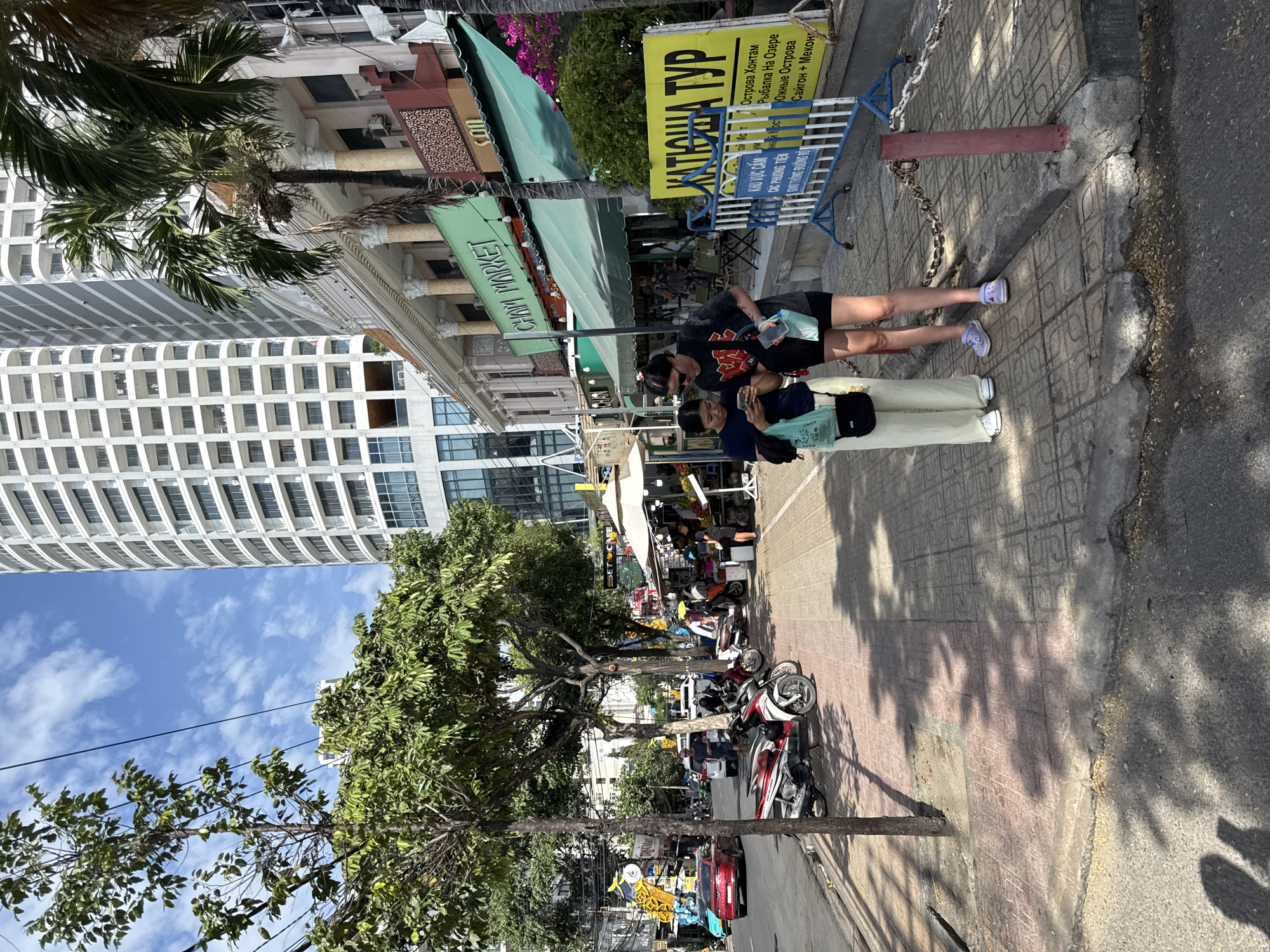 Two young women are standing on a sidewalk in Nha Trang, Vietnam, looking at a smartphone. One woman on the left is wearing a dark t-shirt, light-colored pants, and a dark crossbody bag. The woman on the right is wearing a dark t-shirt with a graphic print, black shorts, and white sneakers. They are standing near a chain fence with a yellow sign in front of a building that appears to be a market or shop. The building has a green awning and columns. Behind them, a street is visible with parked motorcycles and a red car. Tall buildings and palm trees are in the background. The sky is partly cloudy.