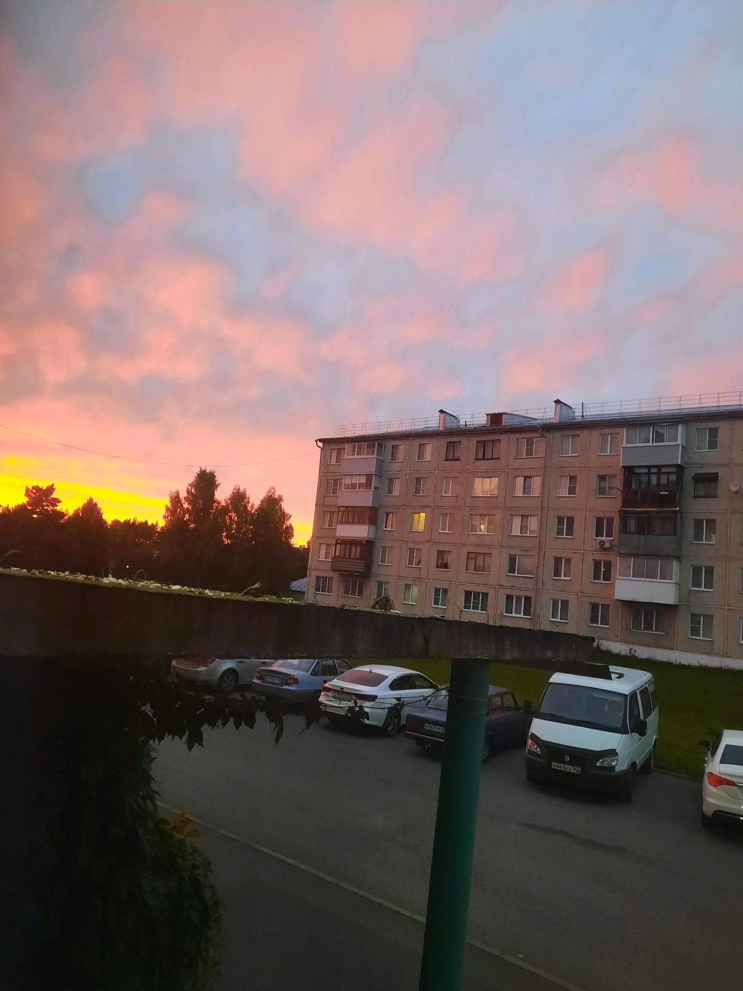 This is a full, outdoor, medium shot of a residential building in Zelenogorsk, Russia, at dusk. The sky is filled with vibrant pink and orange clouds, with a hint of blue. A multi-story apartment building, constructed of concrete panels, dominates the right side of the frame. It features numerous windows, some of which are illuminated, and a recessed balcony on the fourth floor. In front of the building, a row of parked cars is visible, including a white van and several sedans. Trees form a dark silhouette against the colorful sky on the left side of the image. A low, concrete wall runs horizontally across the foreground, with some greenery peeking over it. The overall scene depicts a common urban landscape during sunset.