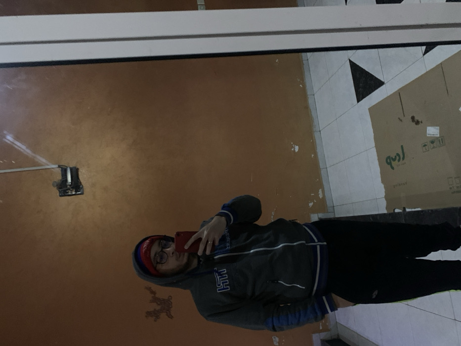 A person is seen taking a mirror selfie indoors. The individual, appearing male, is wearing a gray hooded sweatshirt with blue trim on the cuffs and hem, black pants, a red beanie, and glasses. A ring is visible on their left ring finger. They are holding a red smartphone up to their face, capturing their reflection. The hoodie has what appears to be "MT HT" or similar text printed on the front.
The setting, reflected in the mirror, shows a room with a plain, brownish-orange wall, which exhibits some patches of lighter, possibly peeling paint. An electrical outlet or fixture is visible on this wall, towards the upper left side of the reflection. The floor is tiled, featuring a pattern of white square tiles interspersed with black triangular tiles. A large, light brown cardboard box with some dark stains and mirrored symbols/text (possibly shipping instructions and non-English script) rests on the tiled floor, positioned towards the right side of the frame. A white trim or frame runs horizontally across the top of the image, likely part of the mirror or a doorway. The lighting appears to be indoor artificial light. Given the provided context, the location is Meknes, Morocco. Emotions are not clearly discernible due to the selfie pose and partial obscuring of the face.