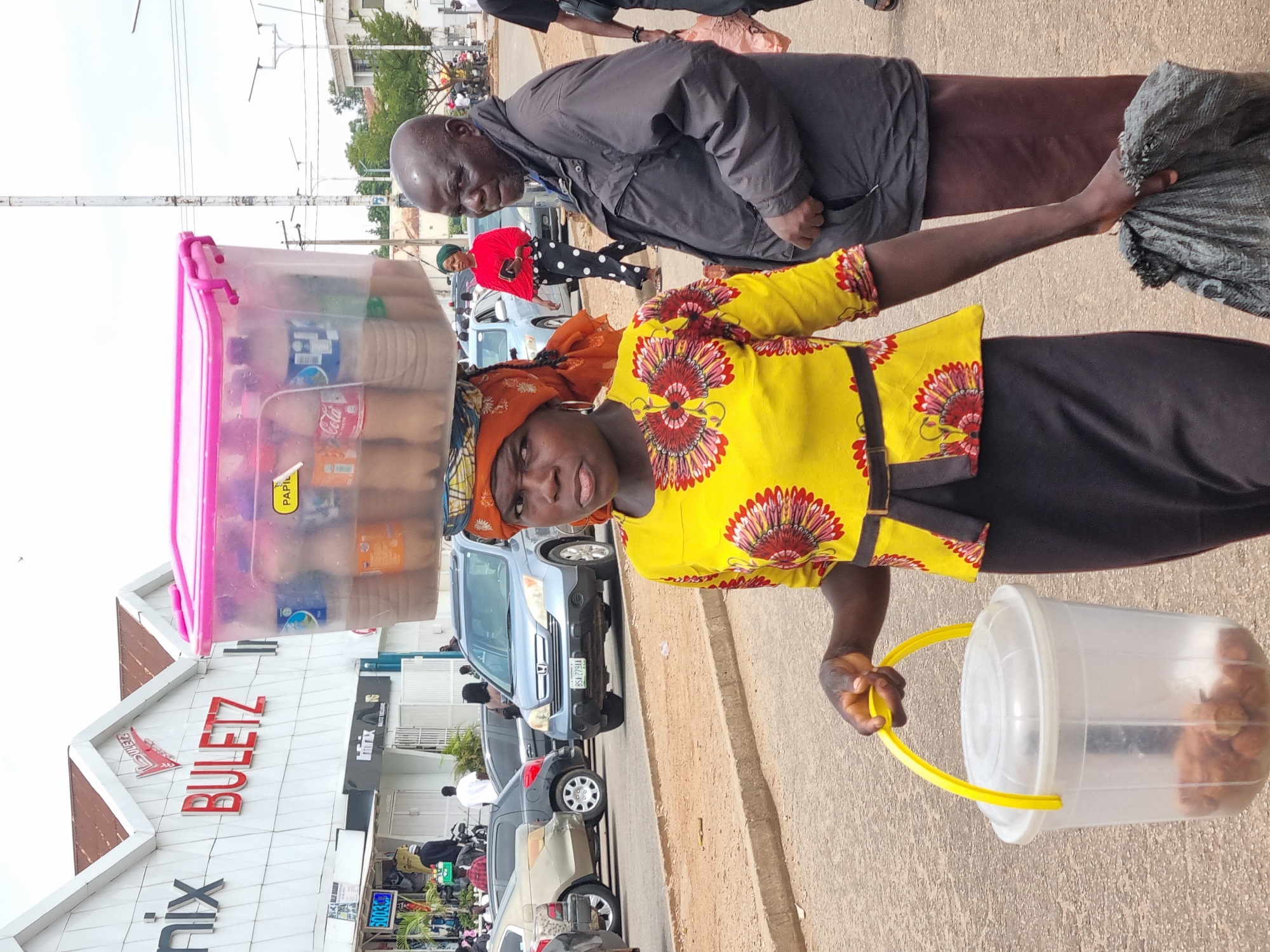 The image shows a street scene in Jos, Nigeria. In the foreground, a woman is walking towards the viewer, carrying a large plastic tub filled with bottles of drinks on her head and holding a yellow-handled clear plastic bucket in her right hand. She is wearing a yellow shirt with a floral print and a black skirt. Beside her, a man is walking away from the viewer, wearing a dark jacket and a brown skirt. In the background, cars are driving on a paved road, and a building with the sign "NIX BULETZ" is visible. Other people are also present on the street. The weather appears to be overcast.
