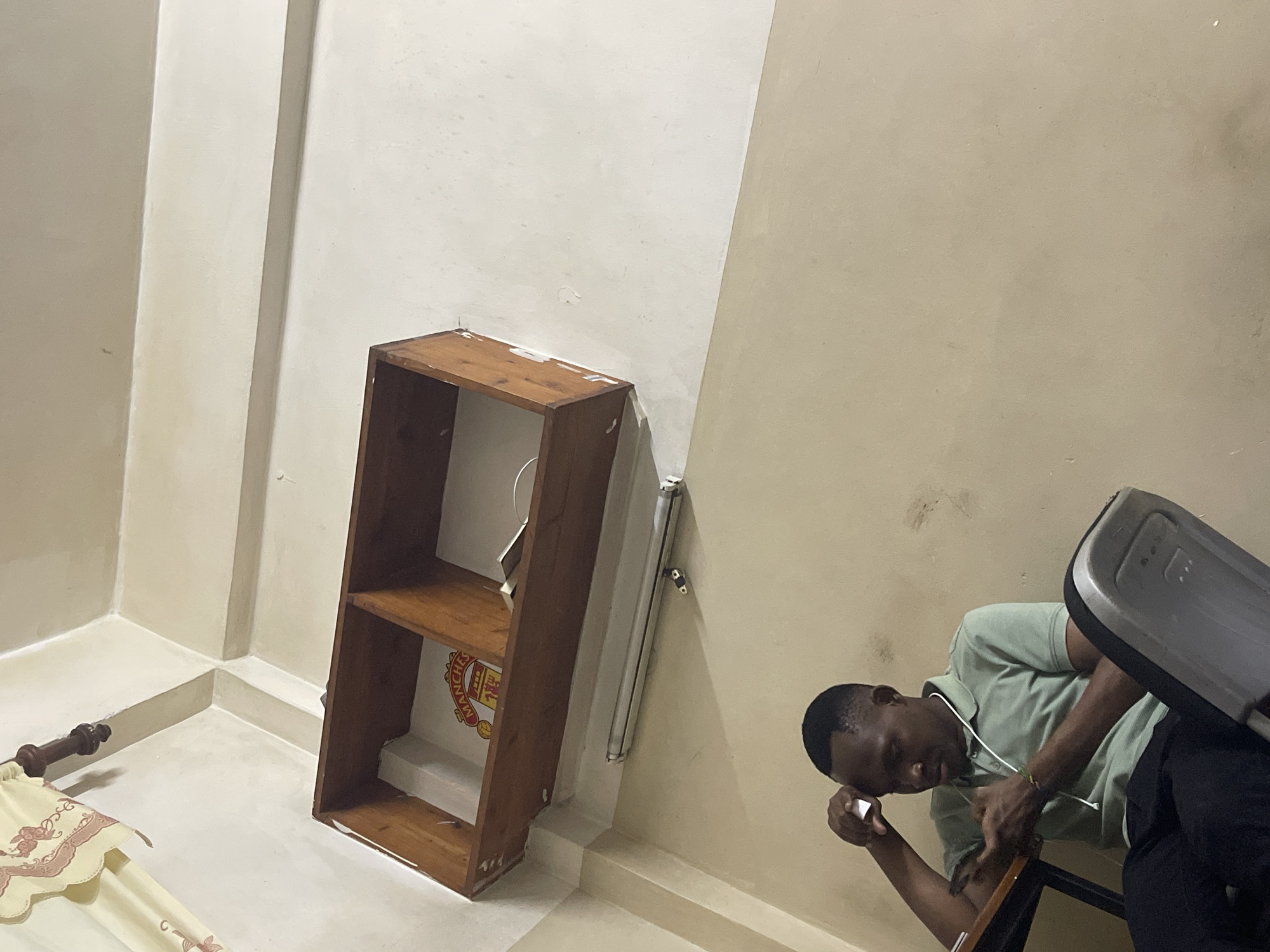 A medium shot captures a room setting. A brown, two-shelf bookcase stands against the wall, bearing a Manchester United logo on its back panel. Adjacent to the bookcase, a fluorescent light fixture is affixed to the wall. In the right portion of the frame, a man with short, dark hair is visible, wearing a light green polo shirt and dark pants. He is seated on a wooden chair with a gray plastic back, seemingly in contemplation, with his left arm resting on the chair. The environment includes light-colored walls and flooring, suggesting an interior space. While the image does not offer explicit information, the Manchester United logo could suggest a fan's personal space or a location where the team is popular. The location in Dar es Salaam, Tanzania cannot be confirmed from the image.