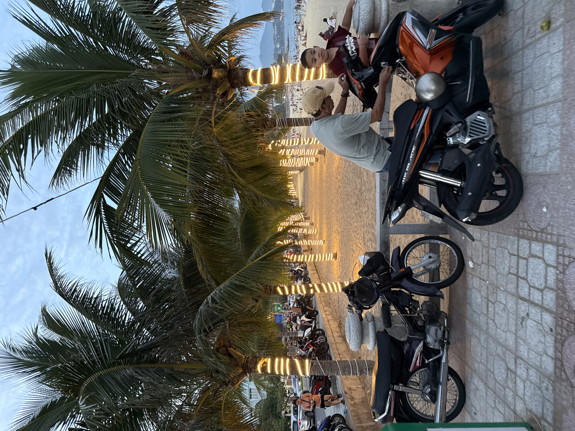 In Nha Trang, Vietnam, along a sandy beach promenade lined with palm trees adorned with string lights, two individuals, an adult male wearing a tan baseball cap and a light green t-shirt, and a young boy in a dark red t-shirt, are interacting with a black and orange motorcycle. The man is leaning over the motorcycle, appearing to adjust something on it, while the boy stands beside him, looking towards the camera. Two other motorcycles are parked nearby on the paved walkway. Beyond the promenade, the beach is populated with numerous people, and the ocean is visible in the background under a twilight sky. Cars are also visible on a street to the left of the palm trees.