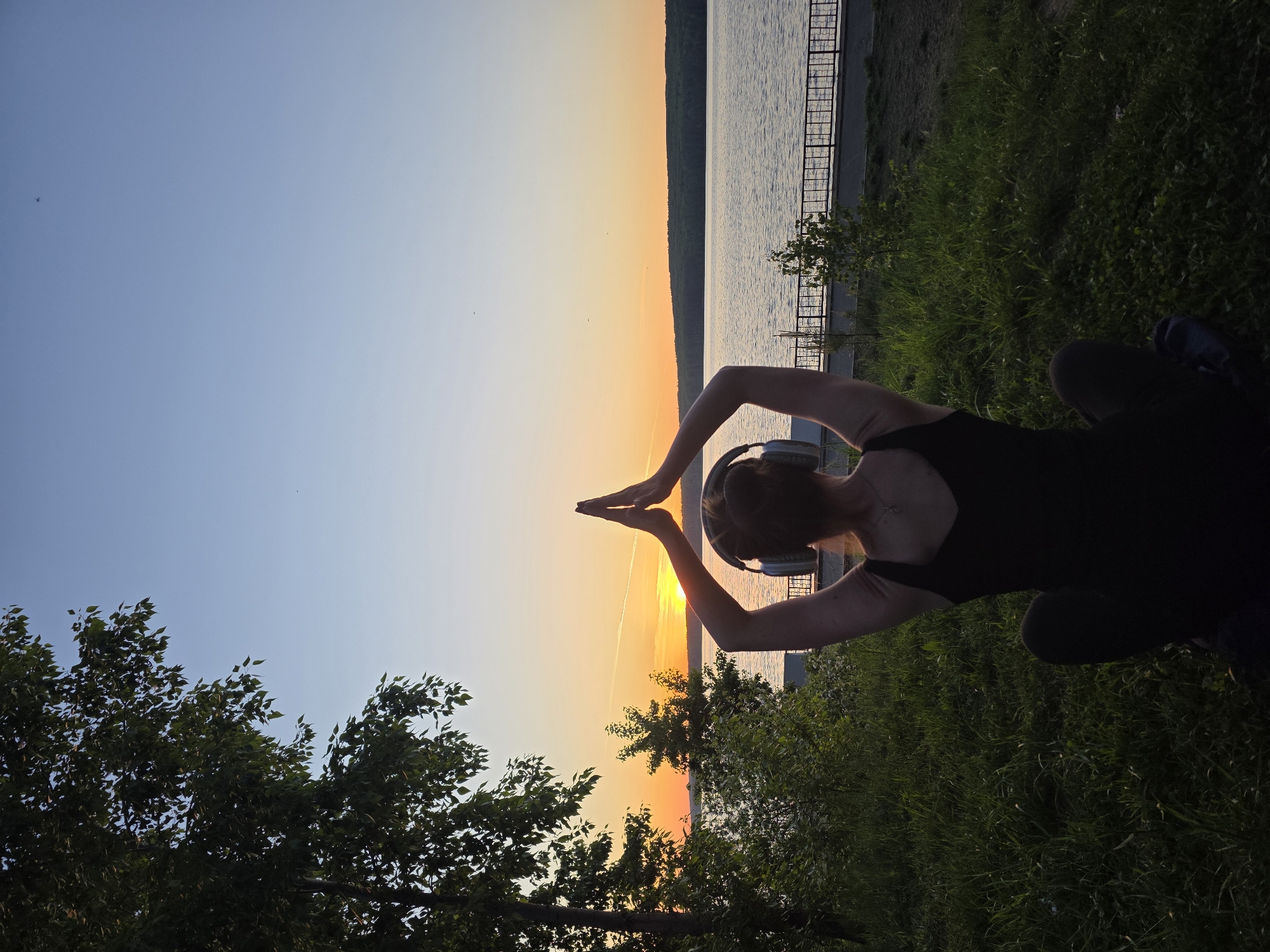 A woman is sitting on a grassy bank, hands raised, making a pyramid shape against the setting sun. She is wearing headphones and is facing the sunset. The background shows a body of water and a shoreline with trees. A fence runs along the bank. The scene is lit by the sunset, casting long shadows. Based on visual cues and location history, this image was likely taken at the city beach of Chaikovsky, Russia.