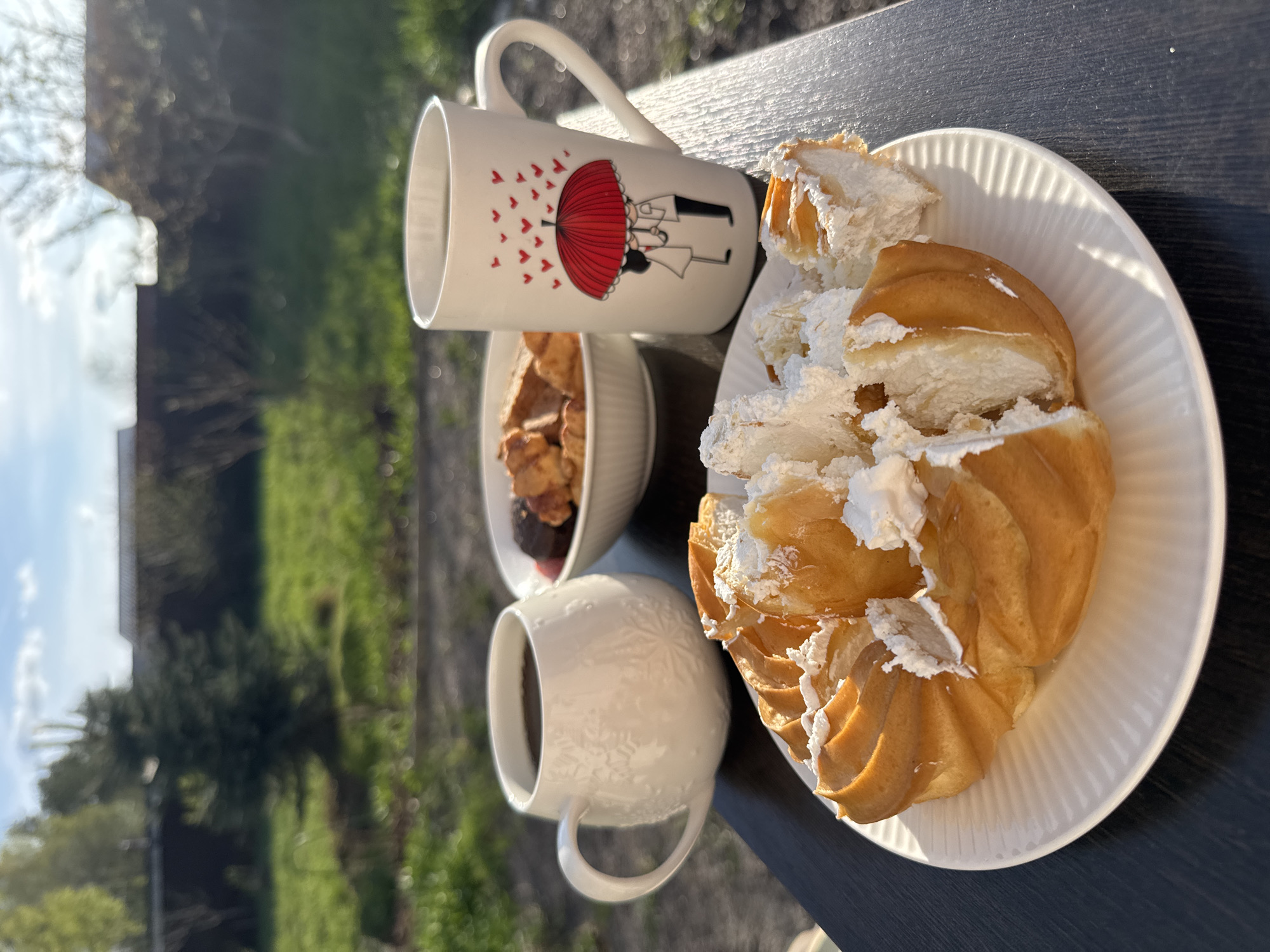 The scene shows a breakfast setting outdoors, likely on a balcony or patio, in Steklyannyi, Russia. There is a dark wooden surface, presumably a table, upon which the food and drinks are placed. Two white ceramic mugs are visible. One mug features a design of a couple under a red umbrella with heart-shaped rain, while the other has an embossed snowflake pattern. A small bowl contains what appears to be granola or baked goods with some fruit. A white plate holds a pastry, possibly a Saint Honoré cake, which has been cut and filled with white cream. The background displays greenery, suggesting a garden or park area. The sky is partially visible with hints of blue. The lighting is bright and sunny, casting distinct shadows.