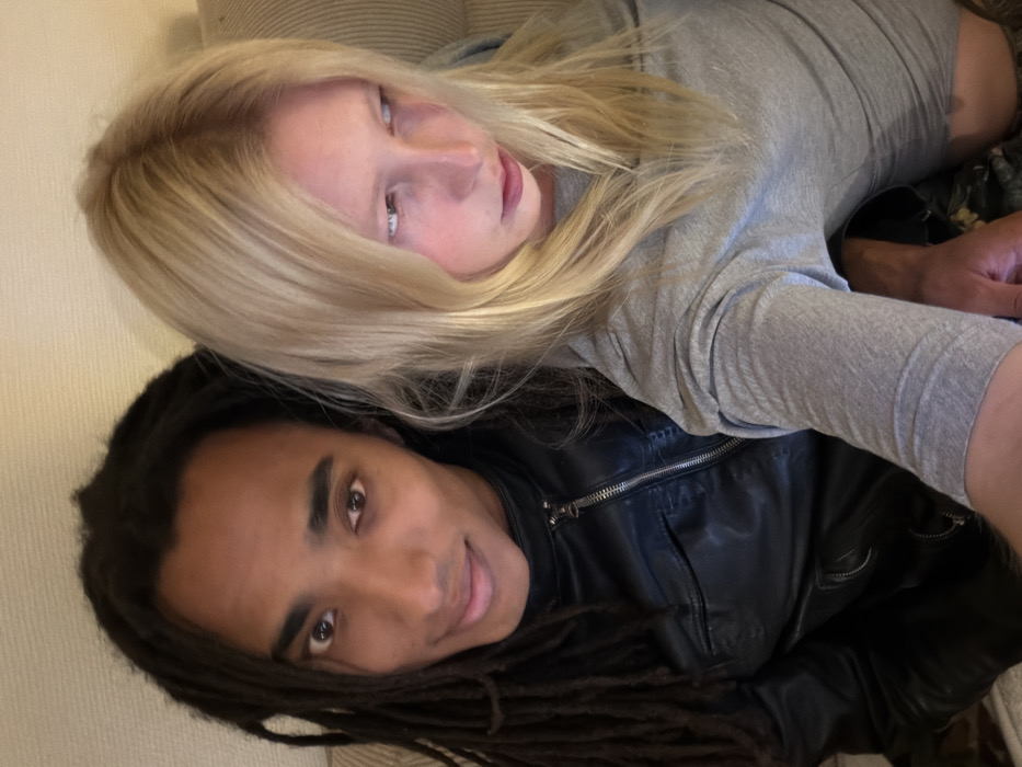The image depicts two individuals in a close-up, indoor setting. The specific location within the city of Paris, France, cannot be determined from the provided visual information, as the background is indistinct and appears to be a plain, light-colored wall or soft furniture.
To the left, a person with dark skin and long dark dreadlocks looks directly at the camera with a slight smile. This individual is wearing a black leather jacket with a prominent silver zipper. To the right, another person with light skin and long blonde hair, showing darker roots, is looking downwards and to the right, away from the camera. This individual is wearing a grey long-sleeved top. Their bodies are positioned in close proximity, suggesting they are seated or reclining together. A hand is partially visible at the bottom right of the frame, possibly holding a mobile device. The lighting is even, suggesting an artificial indoor light source.