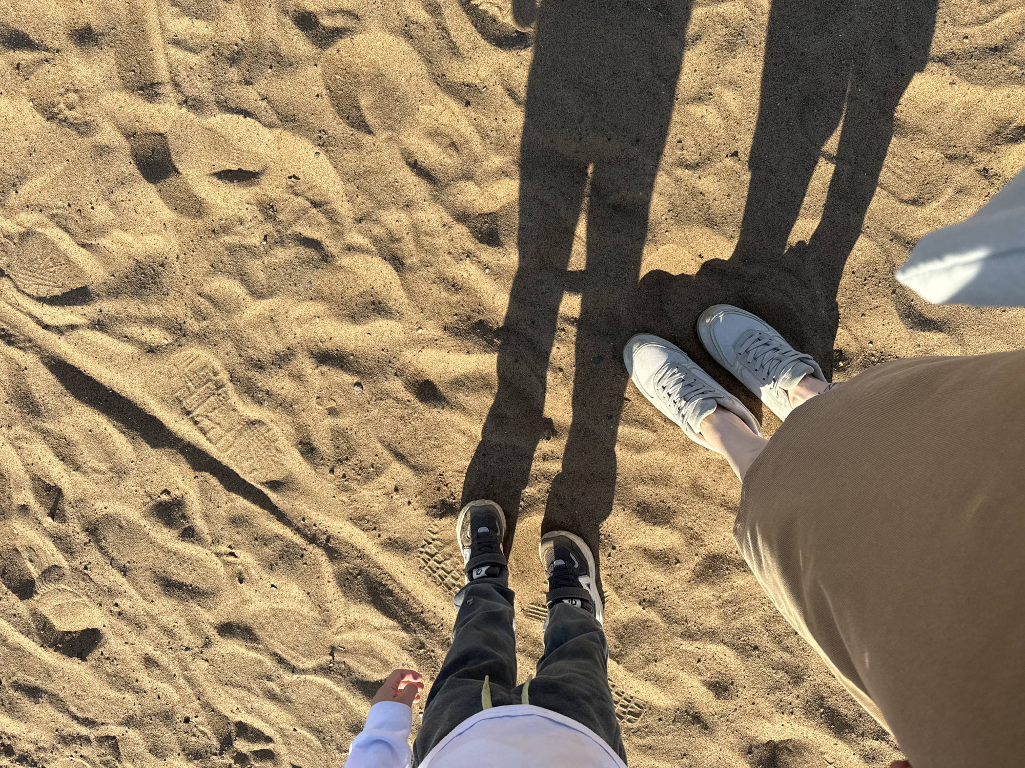 A top-down shot shows two people standing on a sandy surface, likely a beach or a large sandbox. The sand is light brown and appears uneven, with visible footprints and shadows.
The person on the left appears to be a child, wearing dark-colored pants and sneakers with white soles. A small portion of a white shirt is visible. The person on the right is an adult, wearing beige pants and light gray sneakers.
The shadows of both figures are cast on the sand, indicating a light source above and behind them.
The location is not discernible from the image. There is no information in the image that can be used to determine that it was taken in the city of Chaikovsky, Russia.