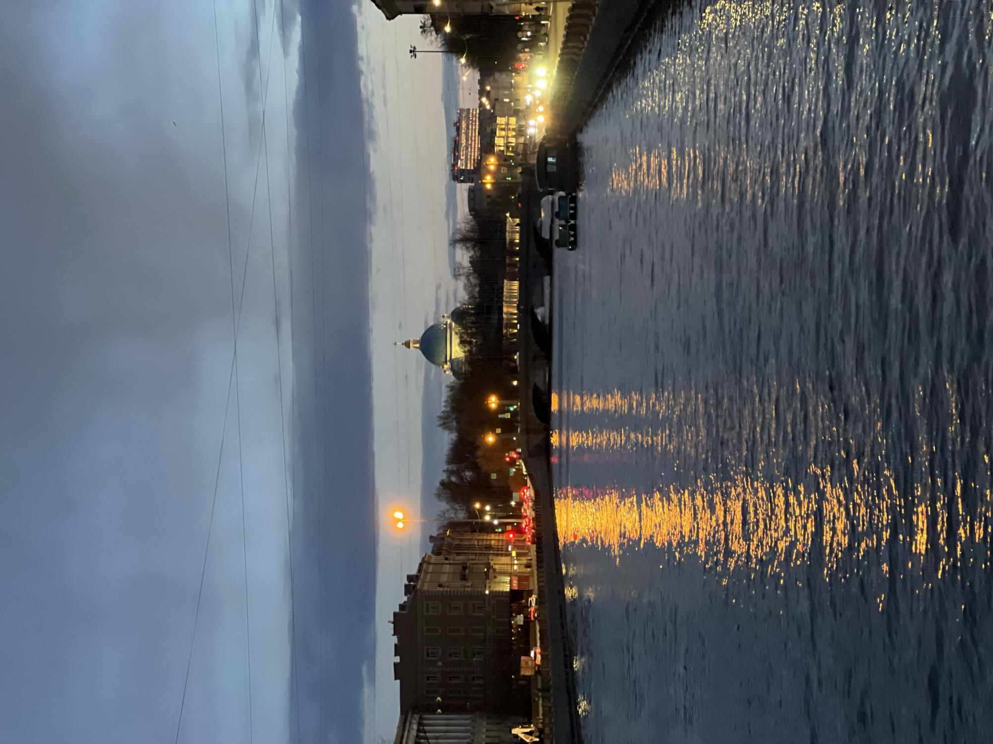 This media file presents a horizontally composed cityscape scene, rotated 90 degrees clockwise in its current display, capturing an evening or night view along a wide river or canal.
In a correctly oriented view, the sky would occupy the upper portion, featuring overcast, dark grey clouds with some lighter patches, traversed by thin power lines. The central part of the horizontal view depicts the city embankment lined with multi-story buildings, illuminated streetlights, and moving traffic. A prominent, light-colored dome with a darker top (possibly blue or green) of a large building or cathedral stands out against the skyline. Trees are visible along the embankment. Car lights, including red taillights and white headlights, create streaks on the road, indicating active traffic.
The lower part of the horizontal view is dominated by the dark, rippled surface of the water, which vividly reflects the yellow and orange artificial lights from the streetlights and buildings. These reflections form shimmering vertical streaks across the water. A few small boats appear to be on the water.
The time of day is late dusk or early night, characterized by the dark sky and widespread artificial illumination. The weather appears overcast with no clear precipitation. The overall atmosphere is that of a calm, bustling city evening. Given the context, this scene is set in Saint Petersburg, Russia, renowned for its numerous waterways and distinctive architecture. No visible text is discernible anywhere in the image.