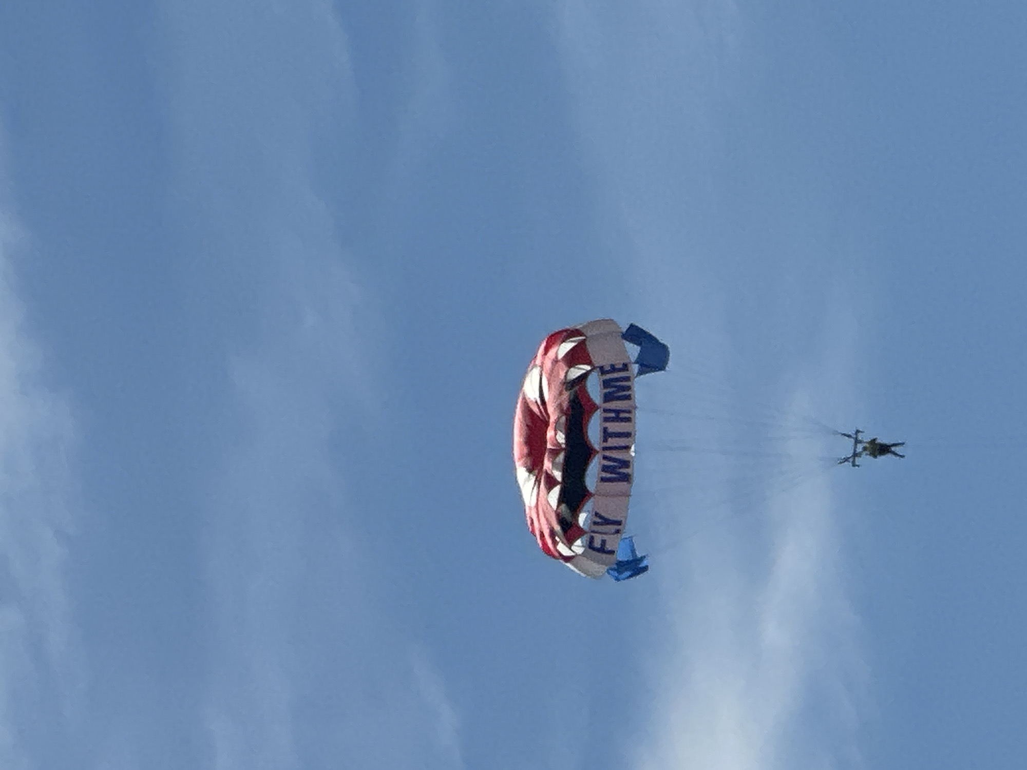 A person is parasailing in a bright blue sky with scattered clouds. The parachute is large and colorful, with red, white, and blue sections. The words "FLY WITH ME" are printed in large white letters across the bottom of the parachute. A single person is suspended below the parachute, attached by lines. The scene appears to be over the water, likely near the coast of Budva, Montenegro, as indicated by the context of the location.