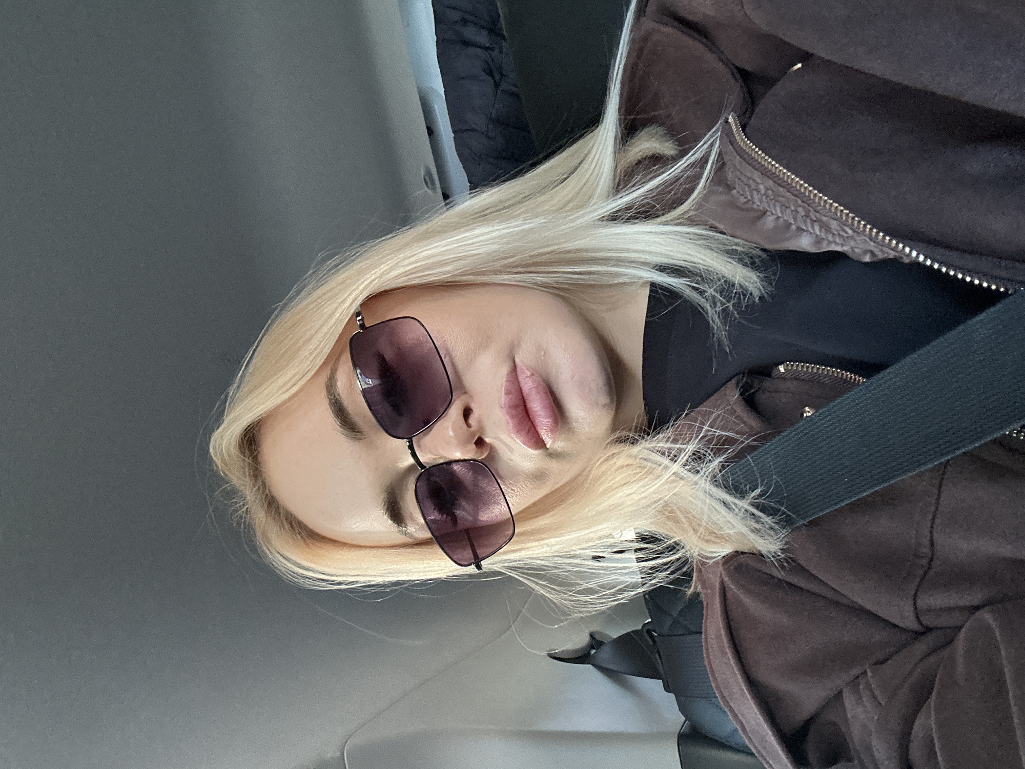 A woman with light blonde hair is sitting in a vehicle. She is wearing sunglasses and a dark-colored top under a brown, zip-up jacket. A seatbelt is fastened across her chest. The interior of the vehicle has a grey panel. The location is unspecified, and there is no information indicating the presence of Megion, Russia.