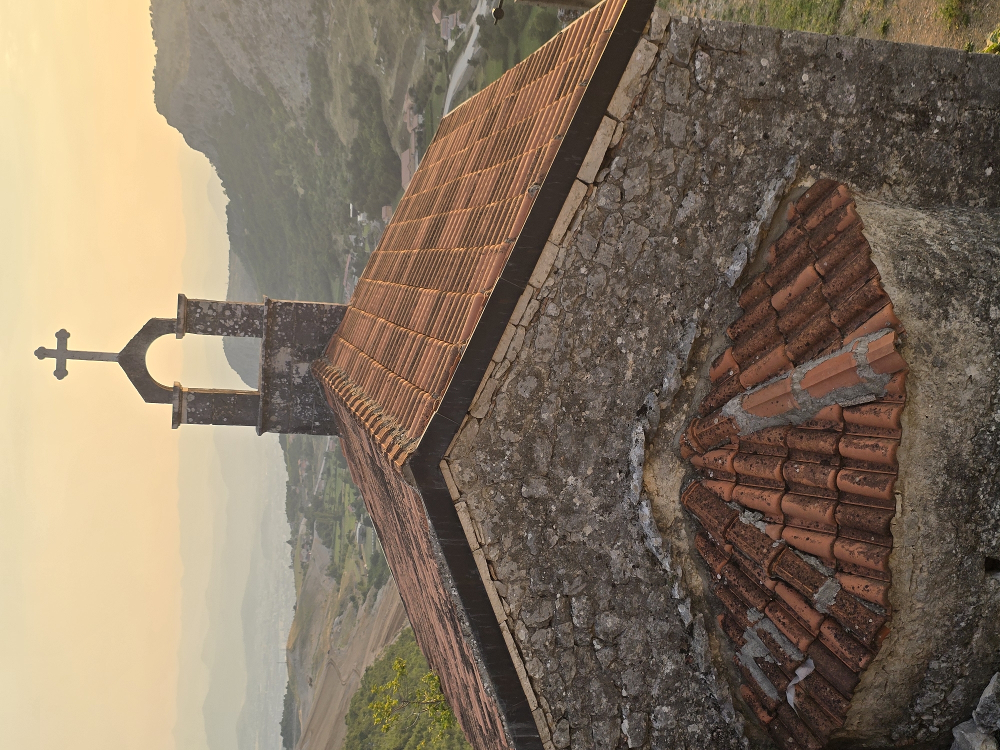 The image captures a close-up view of the roof and bell tower of a small stone church situated on a hillside in Medun, Montenegro. The church features a red tile roof with visible weathering and some areas of missing or dislodged tiles. The stone walls of the church are rough-hewn and appear aged. Above the main roof, a bell tower with an arched opening and a prominent cross is visible against a soft, hazy sky.
The background reveals a rolling landscape with layered mountains fading into the distance. The setting appears to be during either sunrise or sunset, as indicated by the warm, diffused light and the gentle glow on the horizon. Below the church, the terrain slopes downwards, showing some vegetation, fields, and a few scattered buildings of a settlement. A road or path is also visible winding through the landscape. The overall impression is one of a remote, historic religious structure overlooking a rural area.