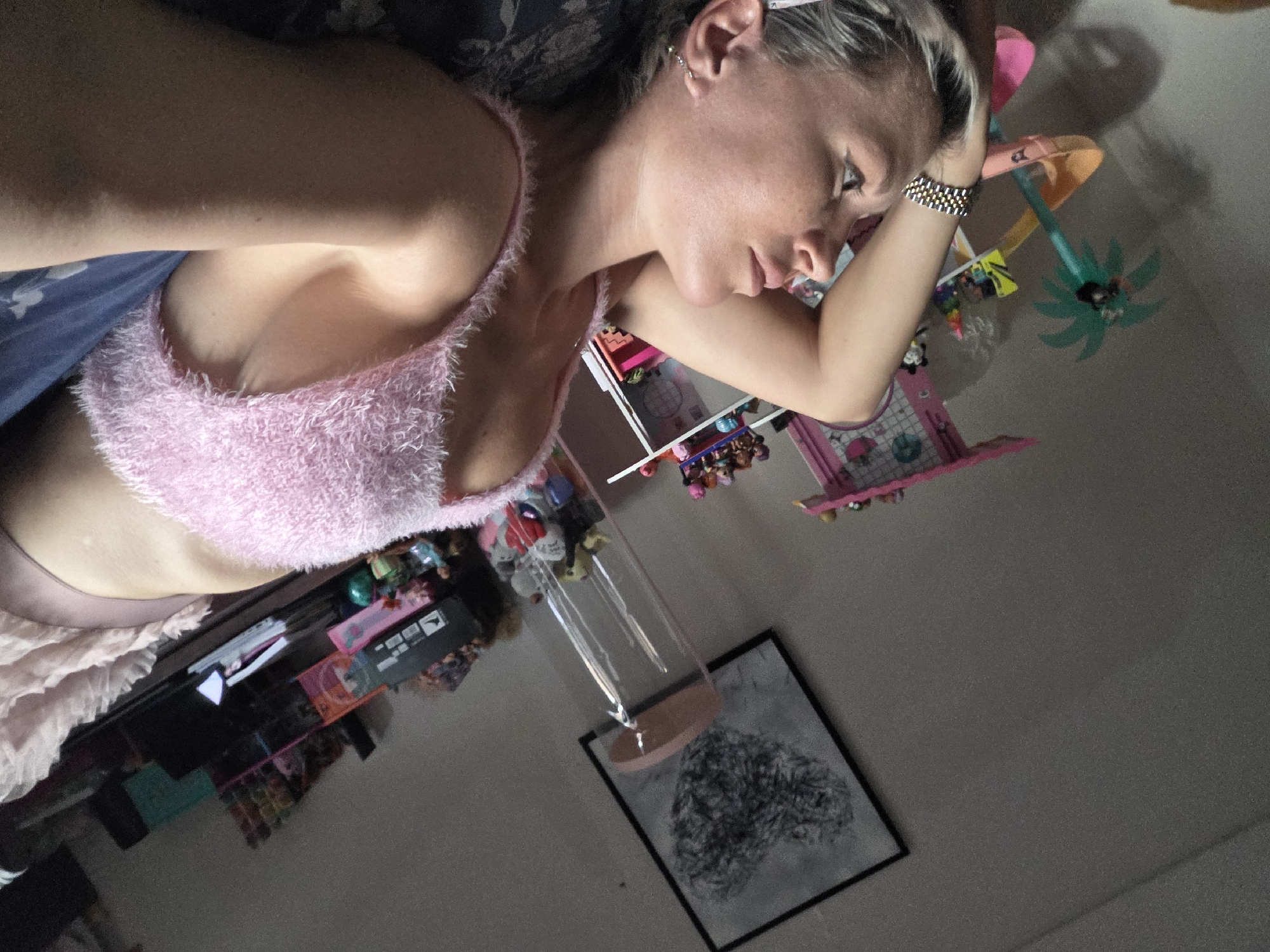 A woman is lying on a bed, looking up at the ceiling. She is wearing a pink fuzzy top and a pink skirt. There are toys and decorations in the background, suggesting a child's room. The room is dimly lit. The overall impression is one of a relaxed and casual setting, possibly in a private residence. The location in North Kuta, Indonesia is not discernible from the image.