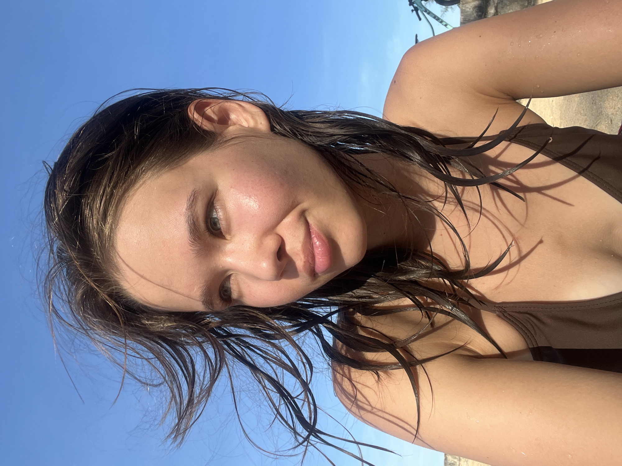 A young woman with wet dark hair and light skin is visible in a close-up, angled selfie shot against a clear blue sky. She is wearing a brown top and appears to be outdoors, possibly near a beach or water source given the wet hair. A bicycle is slightly visible in the upper right corner, suggesting the location may be a park or recreational area. The scene could be in Nha Trang Ward, Vietnam.