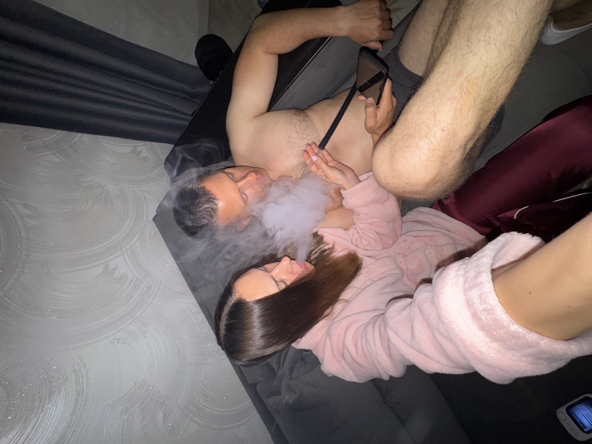 An overhead shot shows a couple lying on a dark gray couch, facing each other. The man is shirtless, wearing dark shorts, and has his left arm bent, resting on the couch. His right hand holds a smartphone, while his left hand is seen in the top left corner of the image. The woman is wearing a light pink fluffy robe and has long dark hair flowing behind her head. A white cloud of smoke billows between their faces, indicating they are likely vaping or smoking a hookah.
The background reveals a light gray wall with subtle, abstract circular patterns, possibly wallpaper, and appears to have glittery specks, suggesting it might be decorative. A dark gray or black curtain is visible on the left side of the image. The lighting is dim, suggesting it might be nighttime or a softly lit indoor setting. No specific location cues beyond the indoor setting are visible, but the context provided indicates the location is Sochi, Russia.
The atmosphere appears relaxed and intimate. The individuals seem to be enjoying each other's company while engaging in a shared activity. No text is visible in the image.