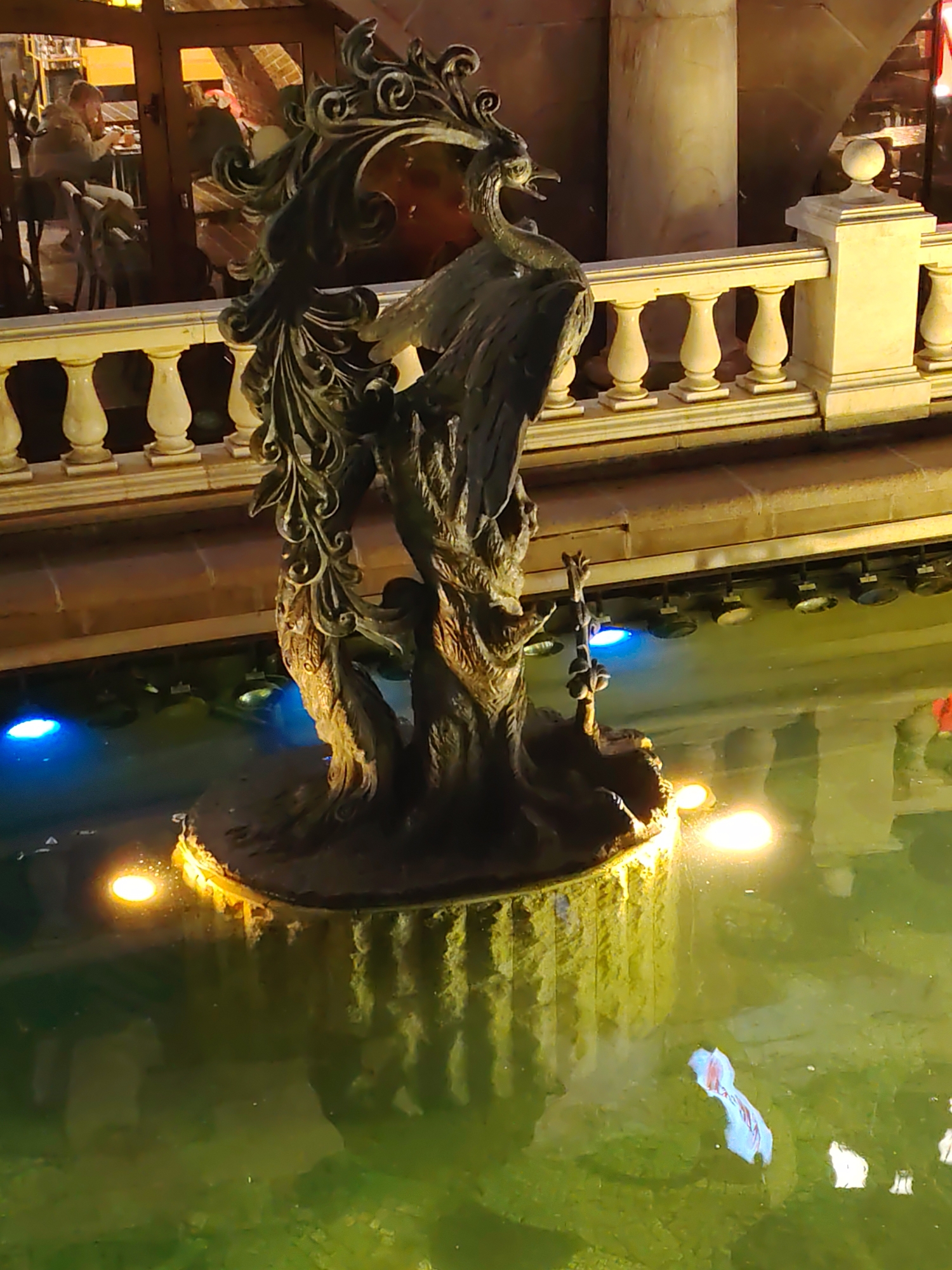 A bronze statue of a phoenix stands in a fountain at night. The statue is dark and ornate, with intricate feather detailing and a beak open as if in mid-cry. The phoenix is positioned on a rocky base, which is surrounded by water. The water is illuminated by several lights, including blue lights submerged in the water and warm-toned lights around the base of the statue, casting reflections on the surface. In the background, a white stone balustrade with decorative posts lines a walkway. Beyond the balustrade, a building with large windows is visible, with some interior lighting and dimly lit figures inside. A partial reflection of a red object is visible in the water near the lower right corner of the frame.
