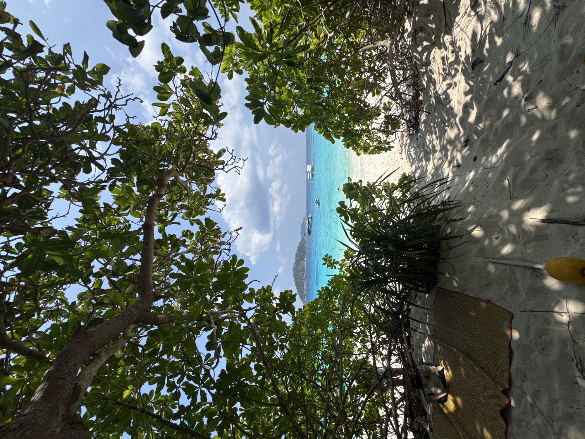 The image captures a view of a beach in Lam Kaen, Thailand. The perspective is from within foliage, with trees framing a path leading to the sand. The turquoise water is visible, with a few boats anchored offshore. The sand is light in color and appears to have been trodden on. To the right, a woven mat rests against the sand, with some light-colored object next to the mat.