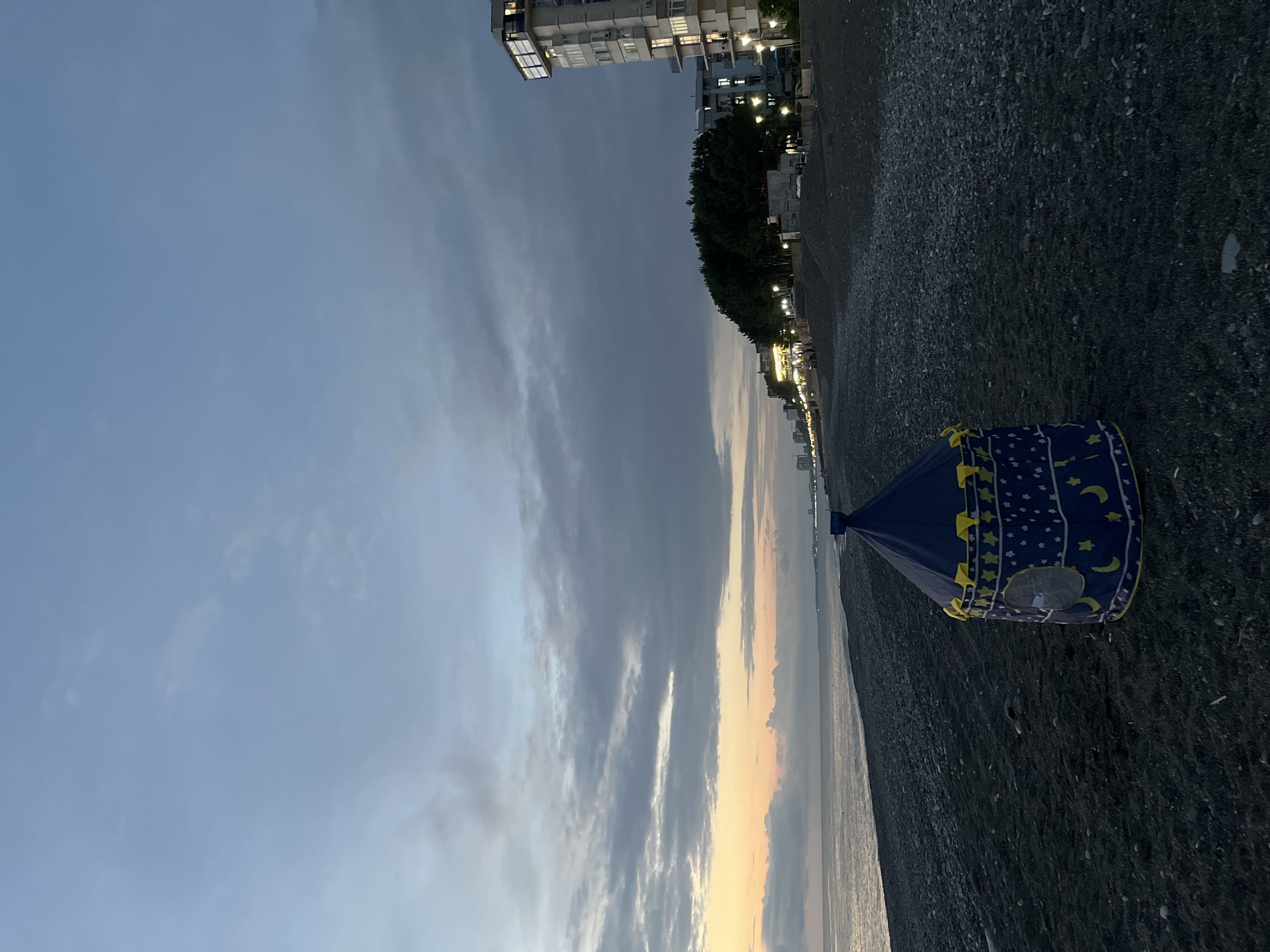 A child's blue tent with yellow star and moon decorations sits on a dark pebble beach facing the Black Sea in Kobuleti, Georgia. The sky is overcast with dramatic clouds tinged with the orange and yellow hues of a setting or rising sun. Along the coastline, a line of trees and buildings with illuminated windows are visible. The sea is calm and reflects the dusky sky. The scene suggests a quiet evening on the coast.