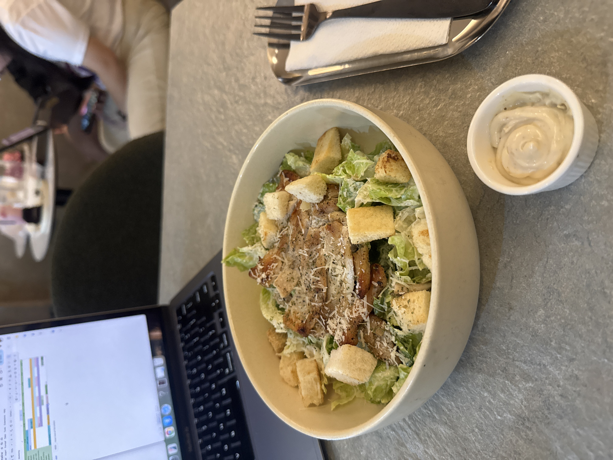 The image shows a Caesar salad in a white bowl on a gray textured surface, next to a small bowl of white dressing and a laptop. A fork and knife rest on a napkin in the upper left. Part of a person is visible in the background. The location is likely a café or restaurant in Davao City, Philippines.