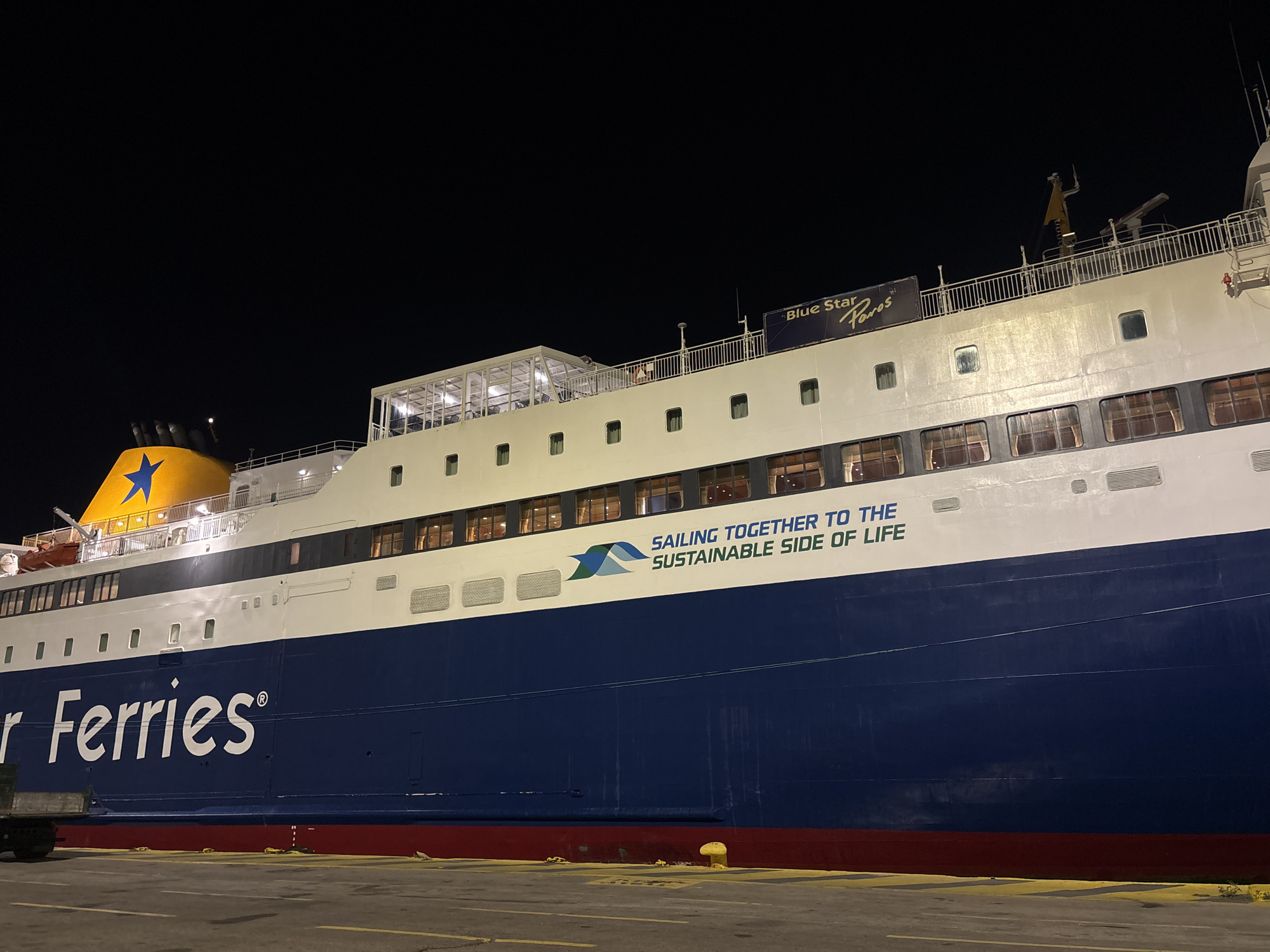 The image shows the side of a large ferry at night. The hull is primarily dark blue, with a white stripe and a red stripe at the bottom. The ferry's name, "r Ferries," is visible in large white letters. Further up, there is text that reads "SAILING TOGETHER TO THE SUSTAINABLE SIDE OF LIFE" accompanied by a green and blue logo. Windows are seen along the upper portion of the ferry. The top of the ferry has a white structure and railings. In the upper left, a yellow smokestack with a blue star is visible. A sign that reads "Blue Star Paros" can be seen on the ship. The photo was taken at night, likely at a port, potentially in Piraeus, Greece, given the presence of Blue Star Ferries, a Greek ferry operator.