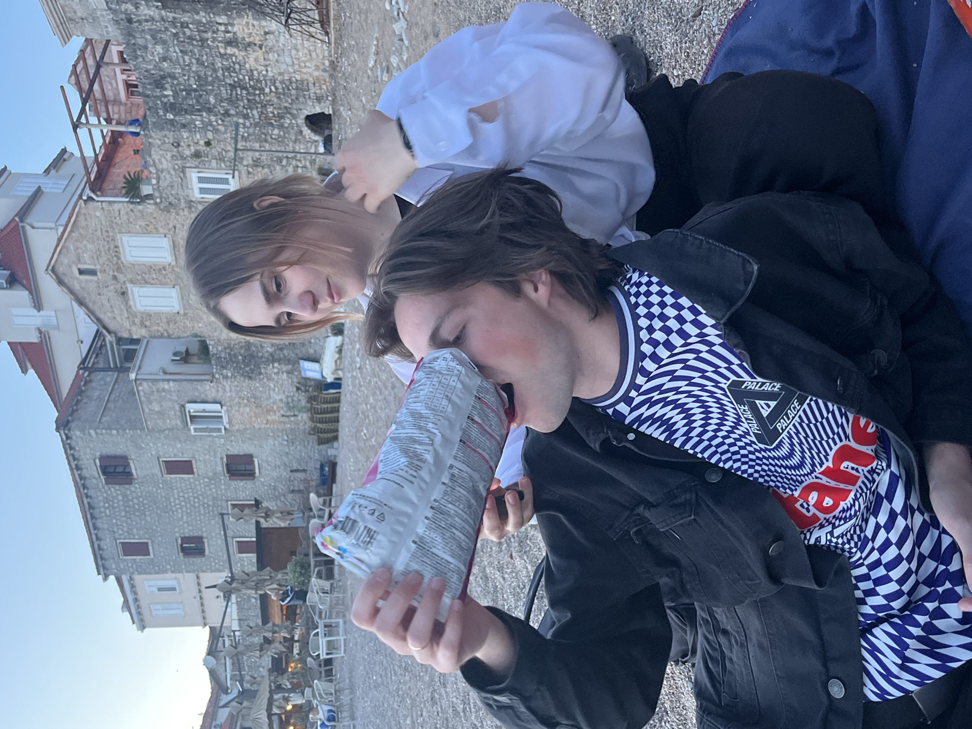 Two individuals are visible in this outdoor scene. One person, positioned closer to the camera, is holding and tilting a snack bag towards their mouth. They are wearing a black jacket over a blue and white checkered patterned shirt with a "PALACE" logo and red writing on the chest. The other person, positioned slightly behind and to the left, is wearing a white shirt.
The setting appears to be a seaside area, potentially a beach, with fine gravel visible on the ground. In the background, a multi-story stone building with numerous windows is present. A portion of the sky is visible. The location is in the city of Unknown, Unknown.