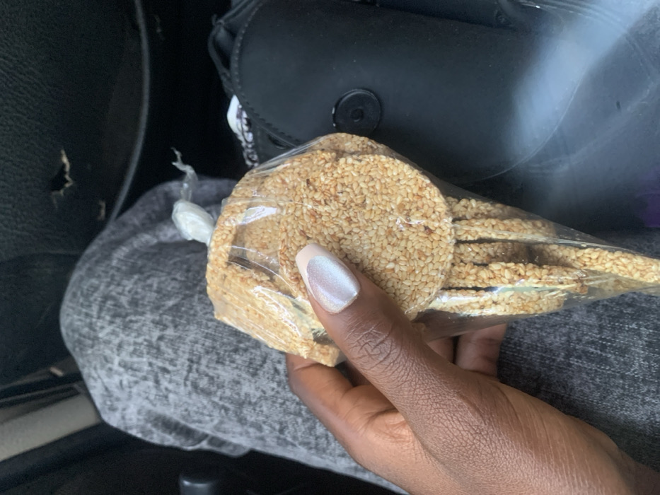 A dark-skinned left hand, featuring manicured nails with an iridescent light-colored polish and lighter tips, holds a transparent plastic bag. The bag contains multiple circular, flat snacks densely coated in sesame seeds, and is sealed at the top with a clear plastic knot. In the background, to the left, a portion of a leg clad in textured light grey fabric trousers is visible. To the right, a black object, consistent with a car seat or vehicle interior panel, shows a tear in its upper section and features a circular black clasp or button. The scene is situated within a vehicle in Rimin Ziem, Nigeria.