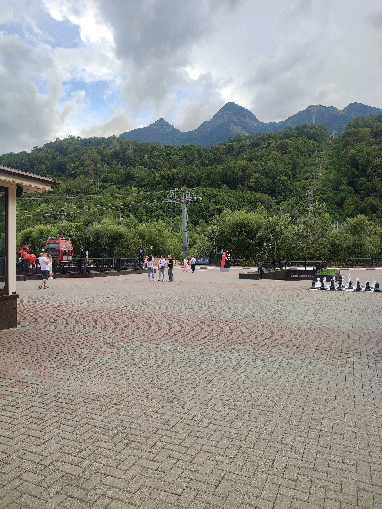 The image captures an outdoor scene in Estosadok, Russia, featuring a paved plaza with a backdrop of lush green mountains and a cloudy sky. In the foreground, a large outdoor chessboard with oversized pieces is set up on the brick-patterned pavement. To the left, a building facade is partially visible. Further into the scene, several people are scattered across the plaza, some walking and others gathered in small groups. A red cable car is parked near the edge of the plaza, with cable car lines extending up the mountainside, indicating a ski resort or mountain transport system. The mountain slopes are densely covered with trees. A sign with "540" is also visible.