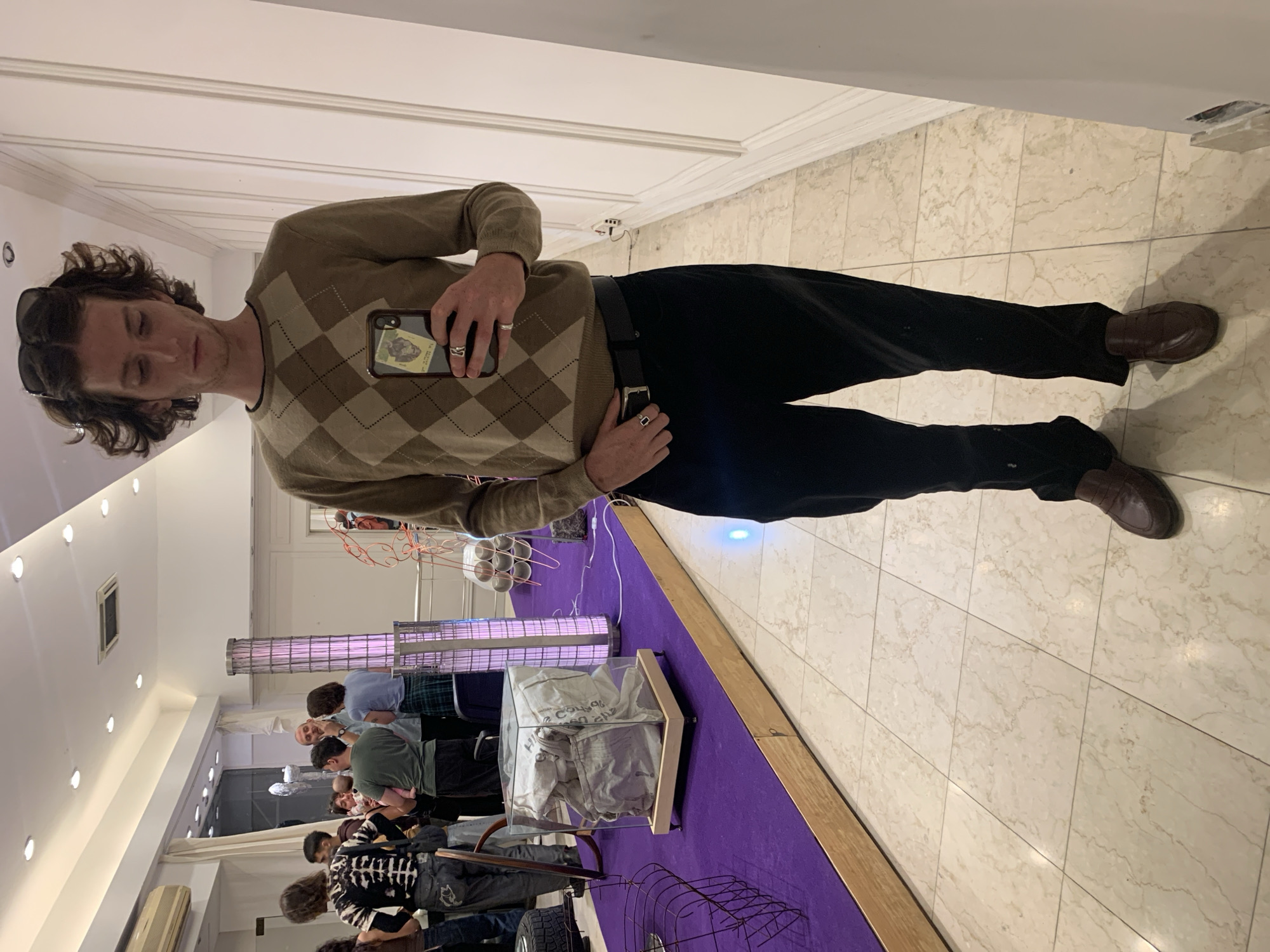 A photograph shows a man standing in an art gallery, possibly in Buenos Aires, Argentina. The man has brown hair, wearing sunglasses on his head, an argyle sweater, black pants, and brown dress shoes. He is holding a smartphone in his left hand, seemingly taking a picture of himself in a mirror. The gallery has a white wall with molding and a tiled floor. In the background, there is an art installation consisting of various objects and a purple carpet. There are people viewing the art in the background. The lighting in the gallery is bright.