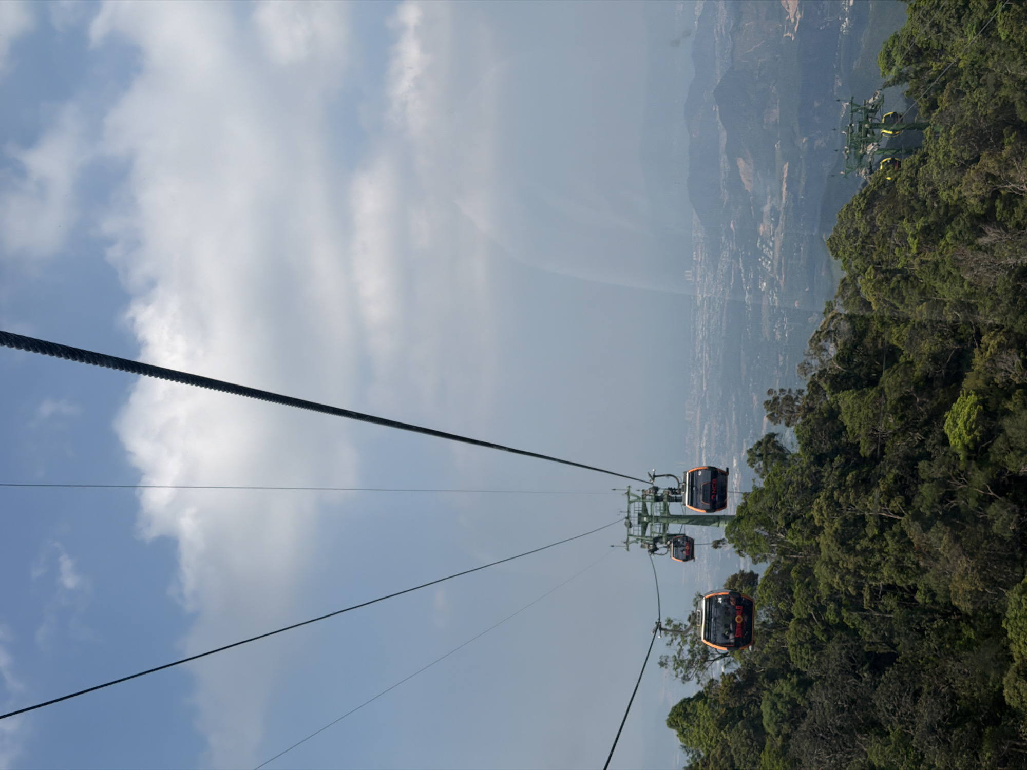 The image depicts a cable car system in Đà Nẵng, Vietnam. The cable cars are suspended on thick, black cables stretching across a blue sky with scattered white clouds. The cars are predominantly orange with black trim. Lush green foliage and trees are visible below and to the right, partially obscuring a hazy cityscape and mountains in the background. Another cable car line with green supports and yellow cars can be seen in the distance.