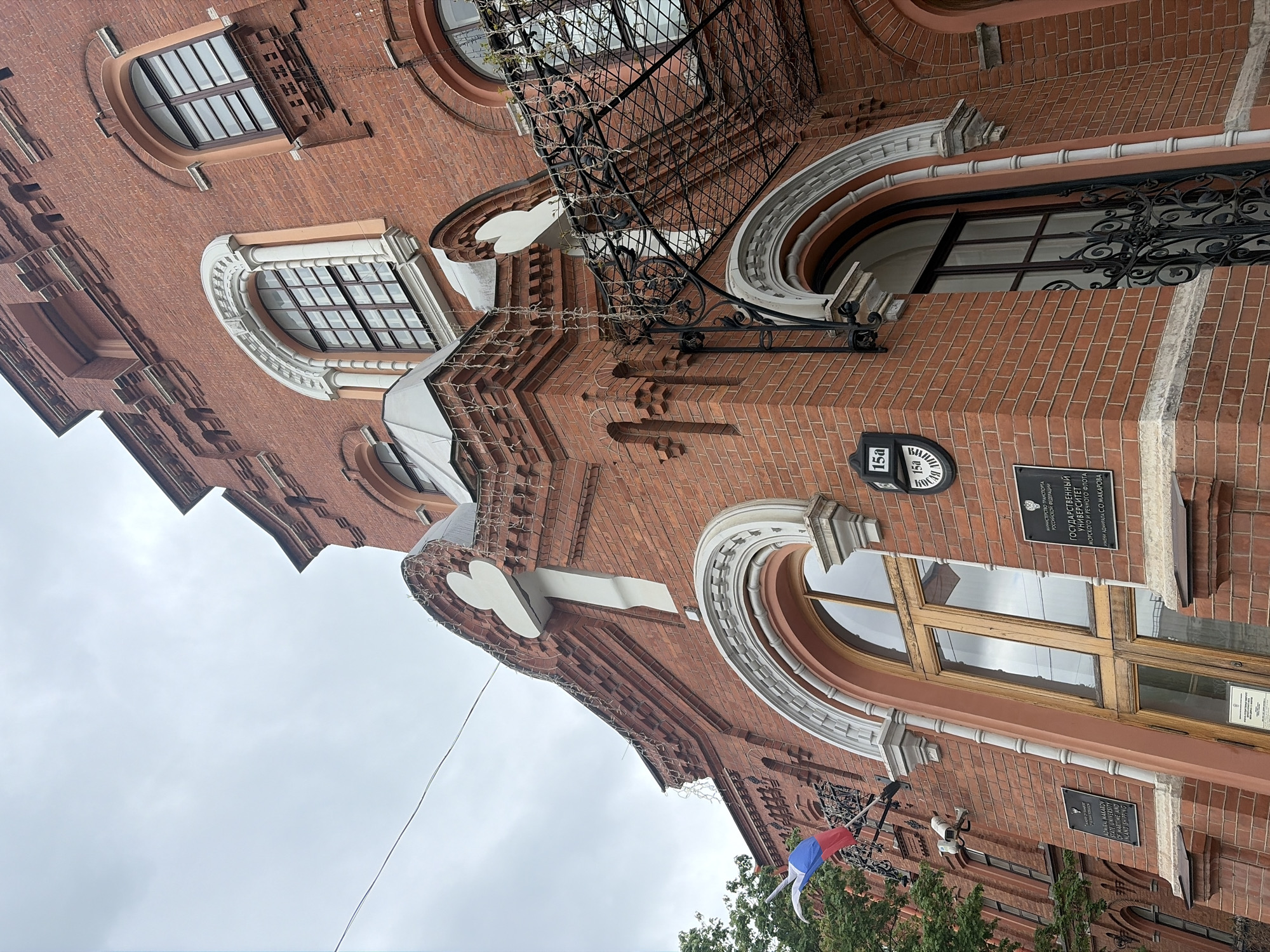 The image shows a red brick building with arched windows, likely located in Saint Petersburg, Russia. Signage on the building identifies it as the Admiral Makarov State University of Maritime and Inland Shipping. A tricolor flag (white, blue, red) is also visible.