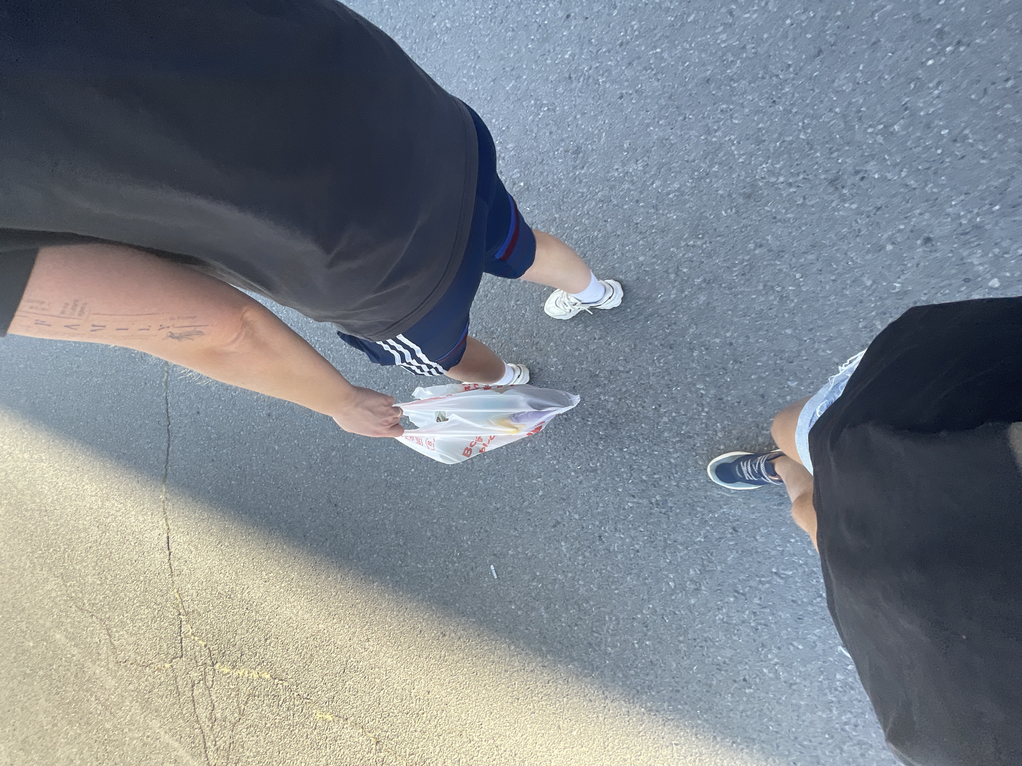Two individuals are walking on a cracked asphalt surface, likely a road or pathway, in what appears to be an urban or suburban setting, possibly in Верхнетемерницкий, Russia. The ground is partially illuminated by sunlight, casting a shadow from an unseen object on the left side of the frame.
The person in the foreground is wearing a black t-shirt and blue shorts. Their legs are positioned as if walking forward, with their right foot clad in a blue and white sneaker and their left foot obscured by their attire.
The person in the mid-ground is also wearing a dark t-shirt and dark blue athletic shorts with white stripes. They are carrying a translucent plastic shopping bag filled with various items. This individual has a tattoo on their left forearm that spells out "FAMILY" vertically. They are wearing white sneakers. Their legs are slightly bent, suggesting they are in motion.
The overall scene captures a mundane moment of people walking outdoors, with a focus on their attire and the simple act of carrying shopping.