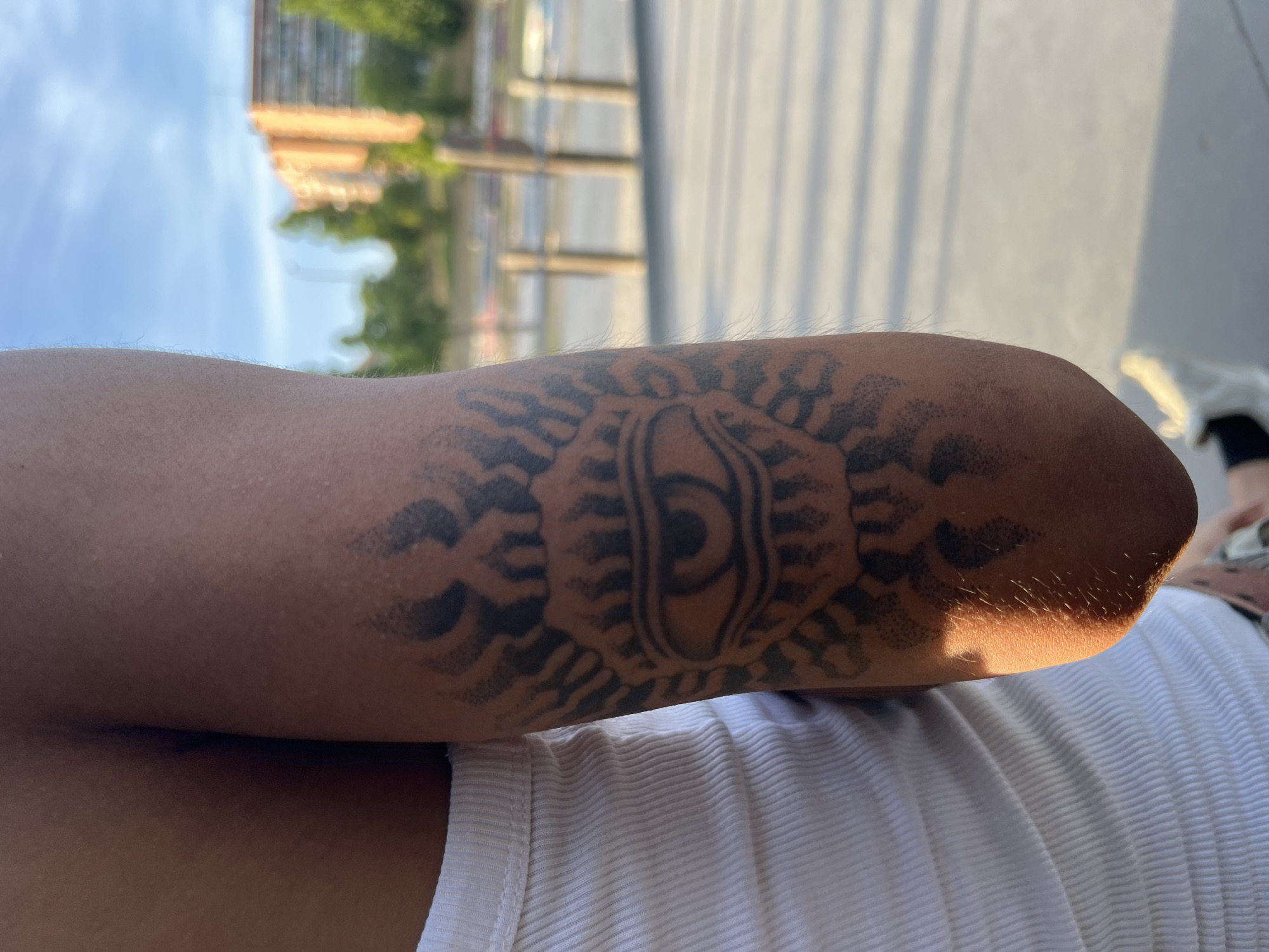 An arm with a black tattoo is resting on a torso clad in a ribbed white tank top. The tattoo is centered on the arm and appears to be a stylized eye design, enclosed by radiating patterns. The scene takes place outdoors, with blurred buildings and trees in the background, indicative of an urban setting. Given the urban setting in Spain, it is not possible to determine the location is Madrid, Spain. The time of day seems to be late afternoon, as indicated by the sunlight and shadows on the arm.
