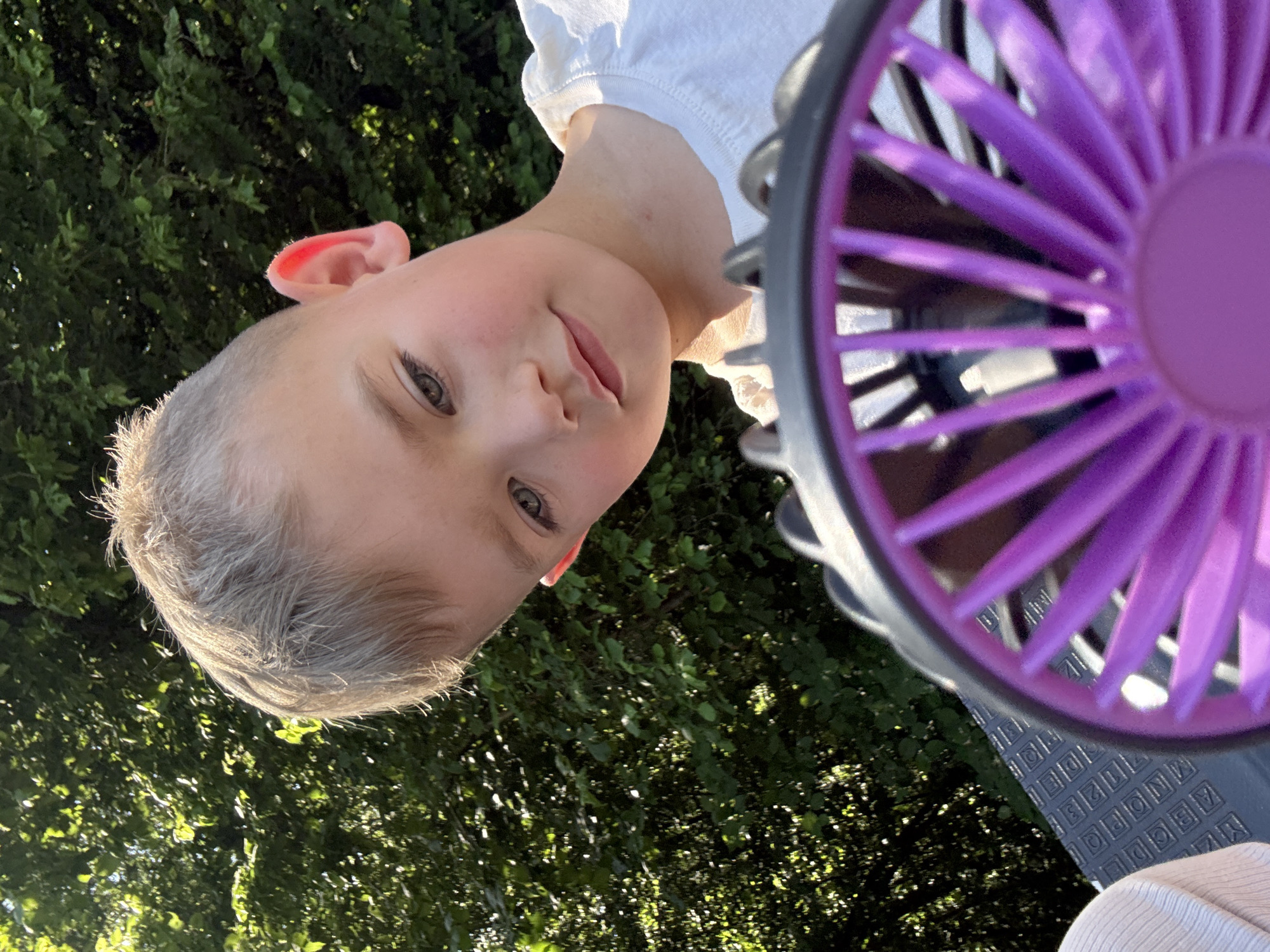 A close-up, eye-level outdoor shot shows a young Caucasian boy looking down and to his left, with a purple and black portable fan in the foreground, partially obscuring his face. The boy has short, light brown hair, fair skin with a light blush on his cheeks, and grey eyes. He is wearing a white t-shirt. His right ear is visible and appears to be flushed pink. Behind him, a dense green background of trees is illuminated by sunlight, creating dappled light effects. The fan has purple blades and a black frame. The boy appears to be in a relaxed pose. The scene is outdoors, possibly a park or garden, during daylight hours. The provided information about the location being Beltershain, Germany, is noted.