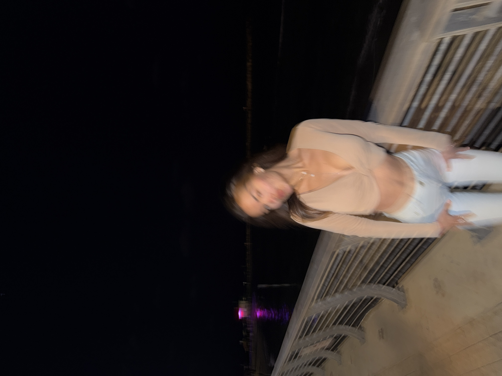 A blurred, nighttime photograph depicts an individual, appearing female, on a promenade overlooking a body of water. The subject, positioned centrally, wears a light-colored long-sleeved crop top with a V-neck and light-colored trousers. Her head is tilted down and to the left, with her eyes closed or partially obscured, and a slight expression on her face. Her long, dark hair is visible.
In the immediate foreground, a metal railing runs horizontally, separating the paved promenade from the water. The railing features multiple horizontal bars and curved supports. The pavement appears to be light-colored tiles or concrete.
Beyond the railing, a dark body of water extends into the background. On the left side of the midground, a structure, likely a building or pier, is prominently illuminated with bright pink and purple lights, which reflect on the water's surface. This illuminated structure and the surrounding environment strongly indicate the location as the promenade adjacent to the Manta Ray restaurant (Dolphinarium Beach) in Tel Aviv, Israel. Faint, distant lights are visible along the horizon line.
The entire image exhibits significant motion blur, particularly affecting the subject, suggesting movement during capture or low-light conditions with a slower shutter speed. The overall lighting is dark, with artificial illumination from distant sources and the prominent pink/purple structure providing the main ambient light.